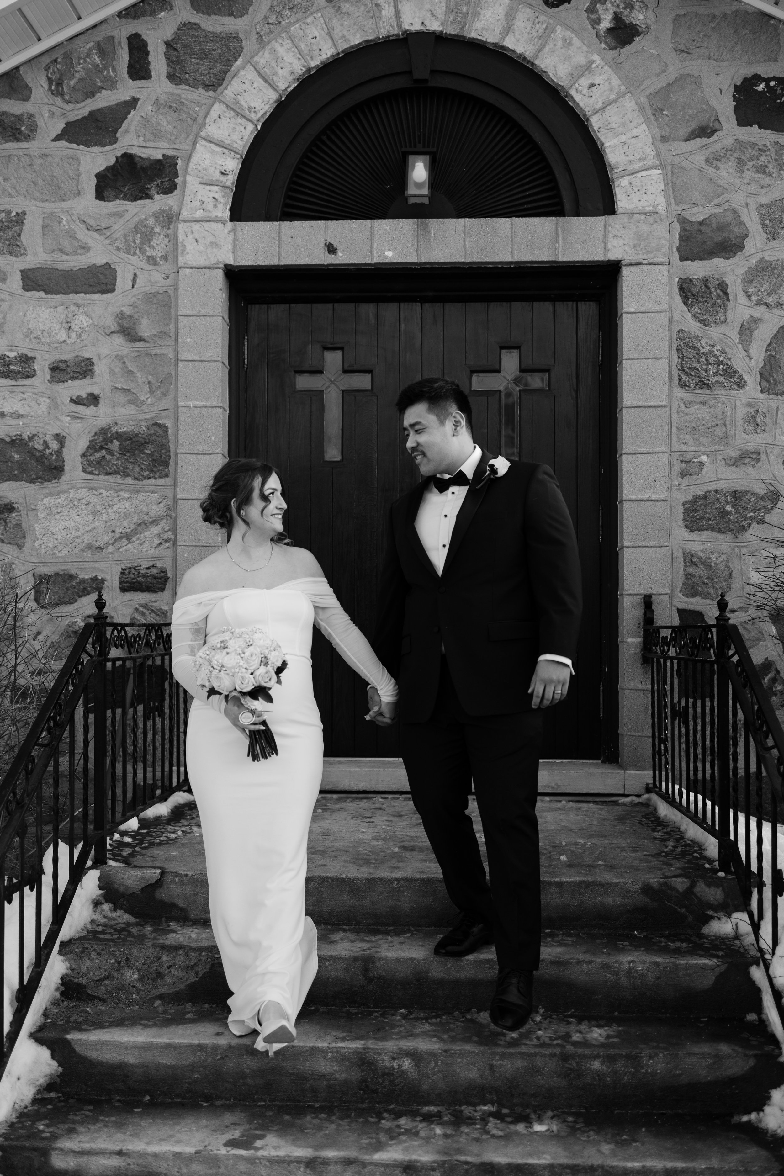 Black and white photo of a bride and groom holding hands on church steps, with stone wall and wooden door with crosses in the background.