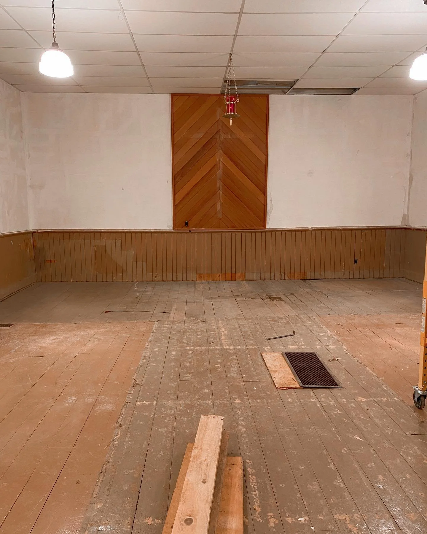 Empty room with unfinished wooden flooring, wood-paneled wall, and unfinished ceiling with some missing tiles. Two ceiling lights are turned on, and there is debris and a vent cover on the floor.