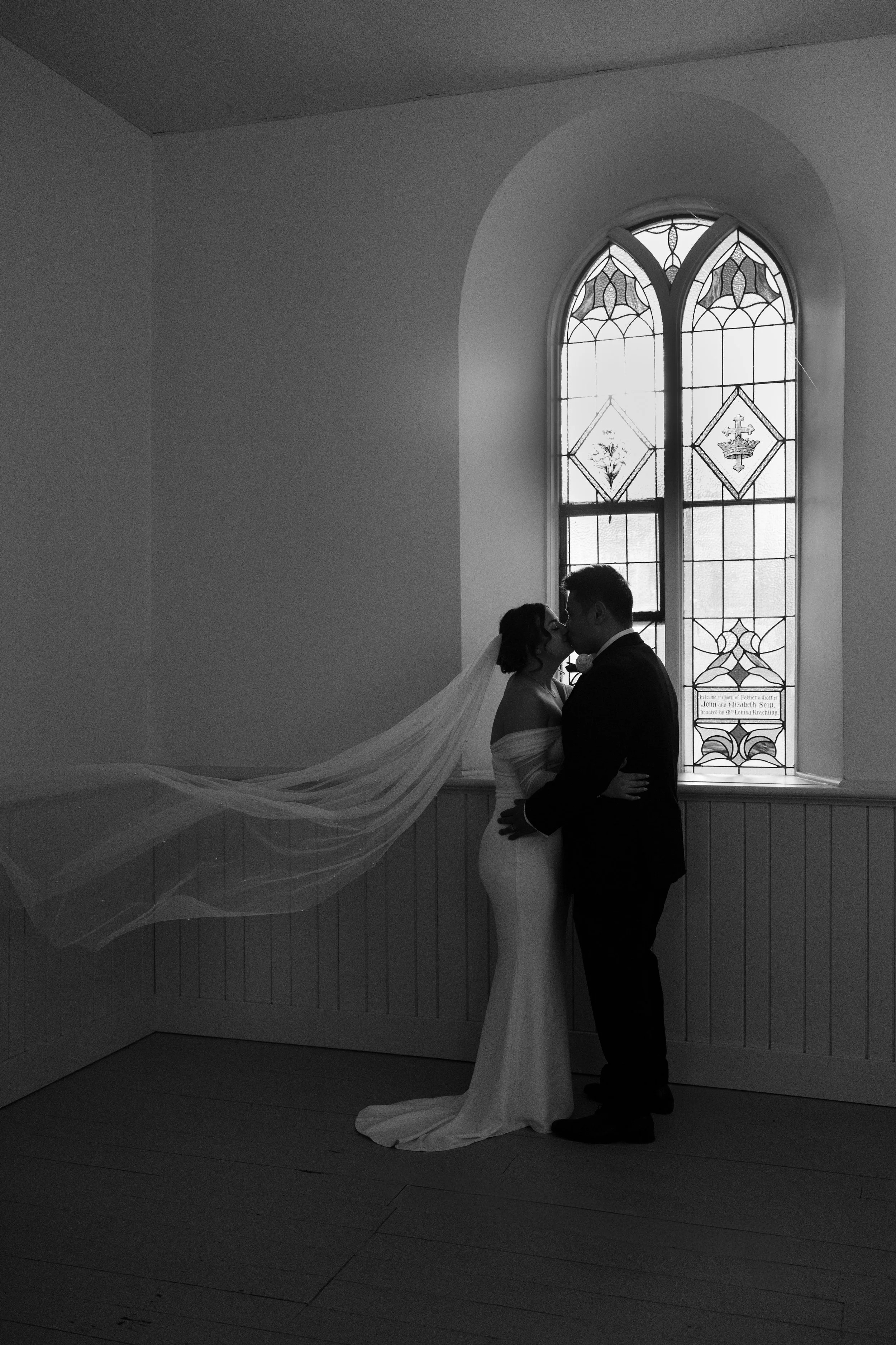 A black-and-white photo of a bride and groom kissing inside a church, standing in front of a stained glass window with decorative patterns.