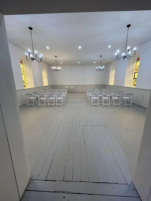 Empty event hall with white walls, wooden floor, white chairs, chandeliers, and stained glass windows
