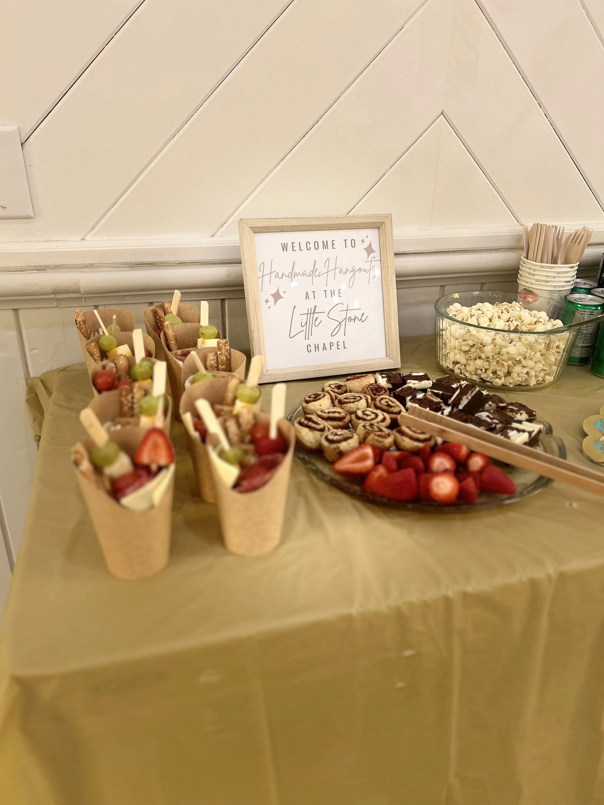Dessert table with fruit-filled ice cream cones, cinnamon rolls, strawberries, chocolate brownies, and popcorn, with a sign welcoming guests to a chapel event.
