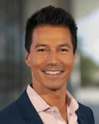 A smiling man in a suit jacket and light-colored shirt, standing outdoors with a blurry background.