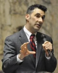 A man wearing a gray suit, white shirt, and red tie speaking into a microphone indoors.