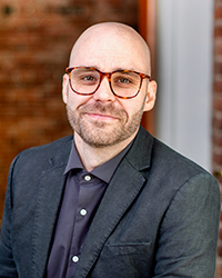A bald man with glasses and a beard, wearing a dark blazer and a dark shirt, smiling at the camera outdoors with a brick building and window in the background.