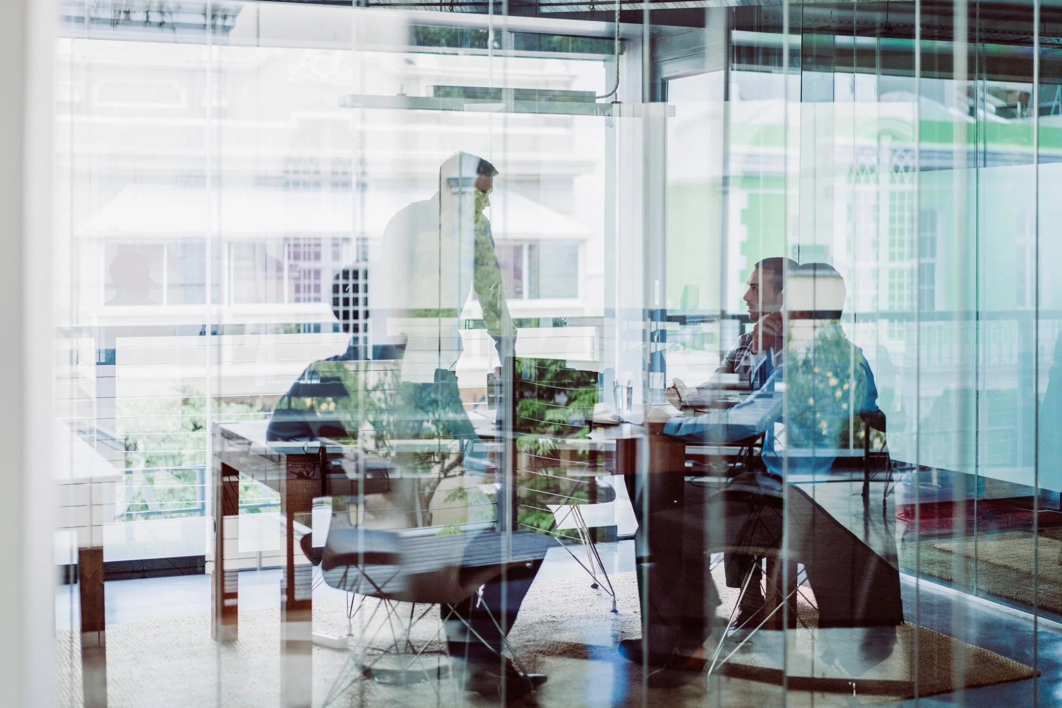 Two men are inside an office, seen through glass walls. One man is standing, leaning over the desk, and the other is seated, looking at the standing man.