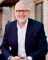 A smiling older man with gray hair and glasses, wearing a dark blazer and white shirt, standing outdoors in front of a building with large windows.