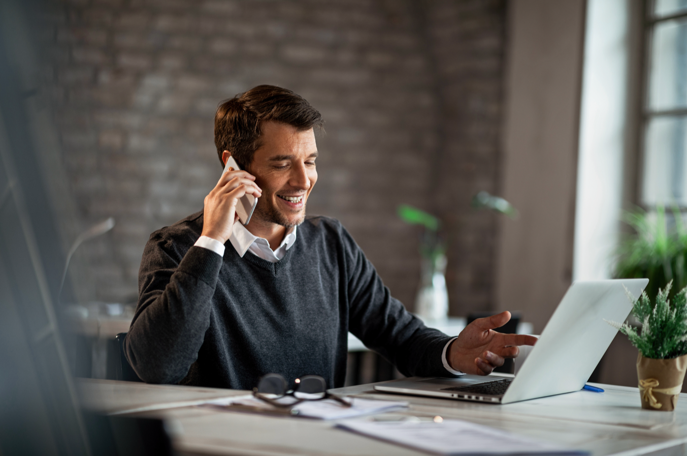A man smiling and talking on his cellphone while working on a laptop at a desk in a modern office.