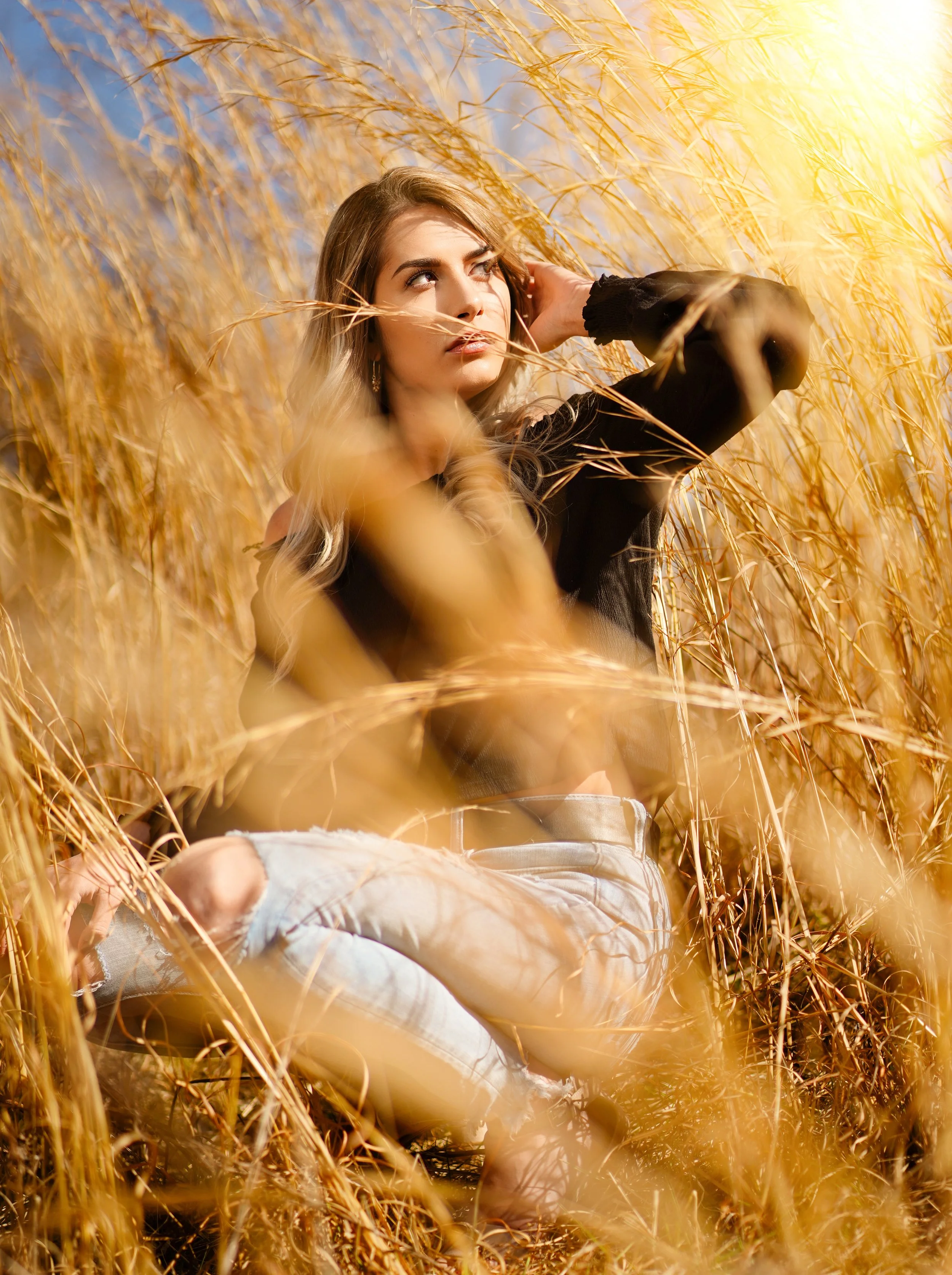 A young woman with long blonde hair sitting in a golden wheat field, wearing torn jeans and a black off-shoulder top, with the sun shining brightly on a clear blue sky.