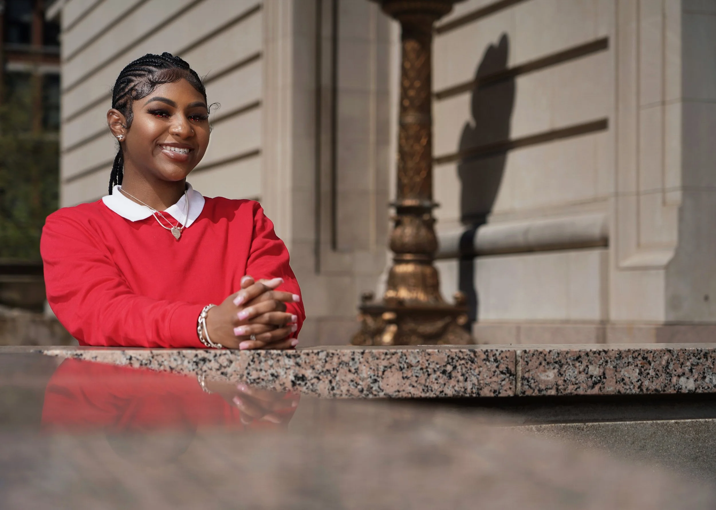 A young woman with dark braided hair, wearing a red sweater over a white collared shirt, smiling with her hands clasped on a granite surface outside a stone building.