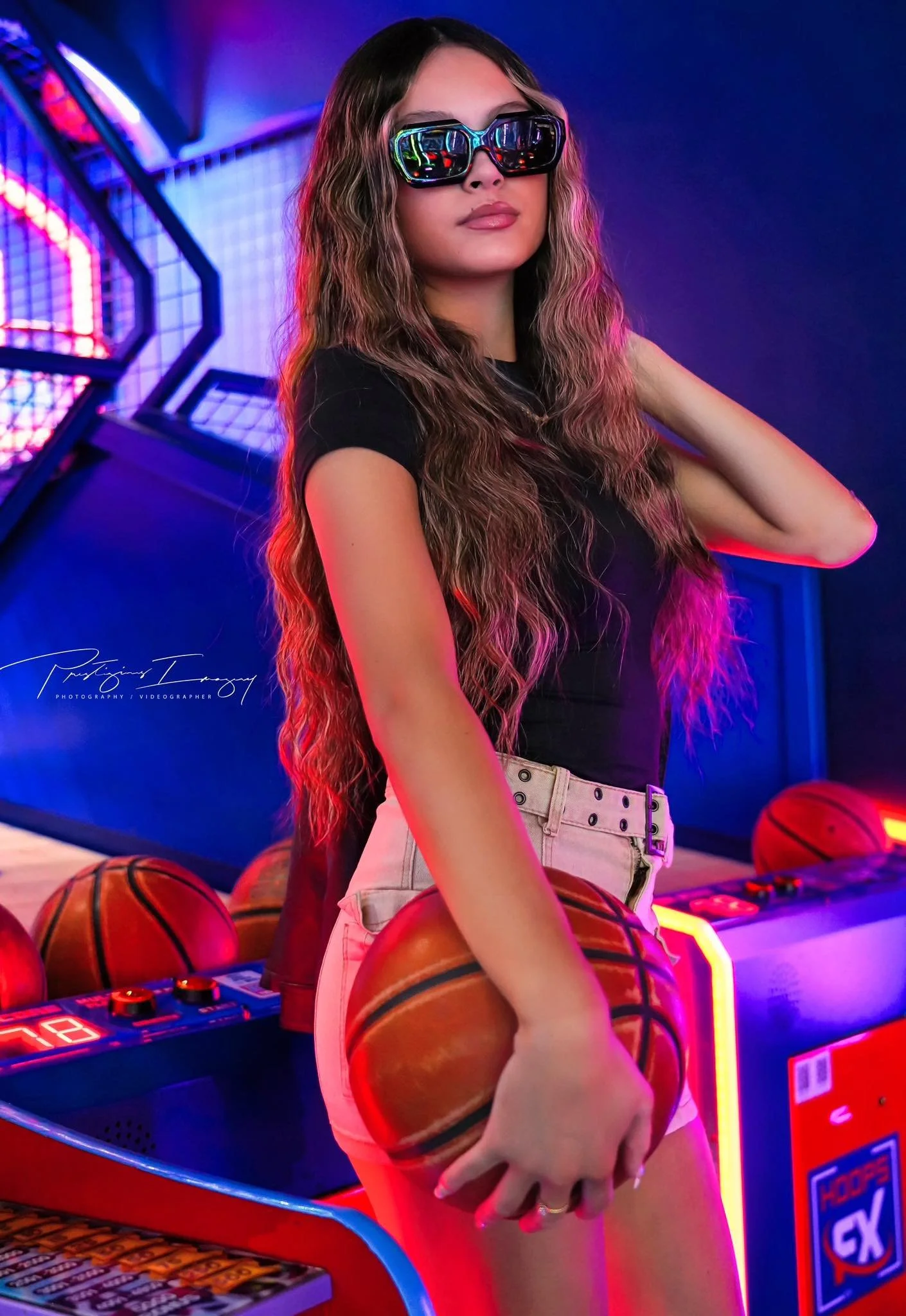 A young woman with long, wavy hair wearing sunglasses, a black t-shirt, and light-colored shorts, holding a basketball in a neon-lit arcade setting with more basketballs and arcade machines in the background.