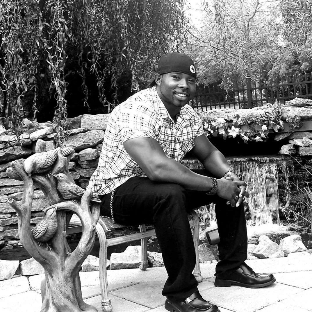 A smiling man in a plaid shirt, black jeans, and boots sitting on a decorative bench with a tree design, in front of a small waterfall and garden, in black and white.