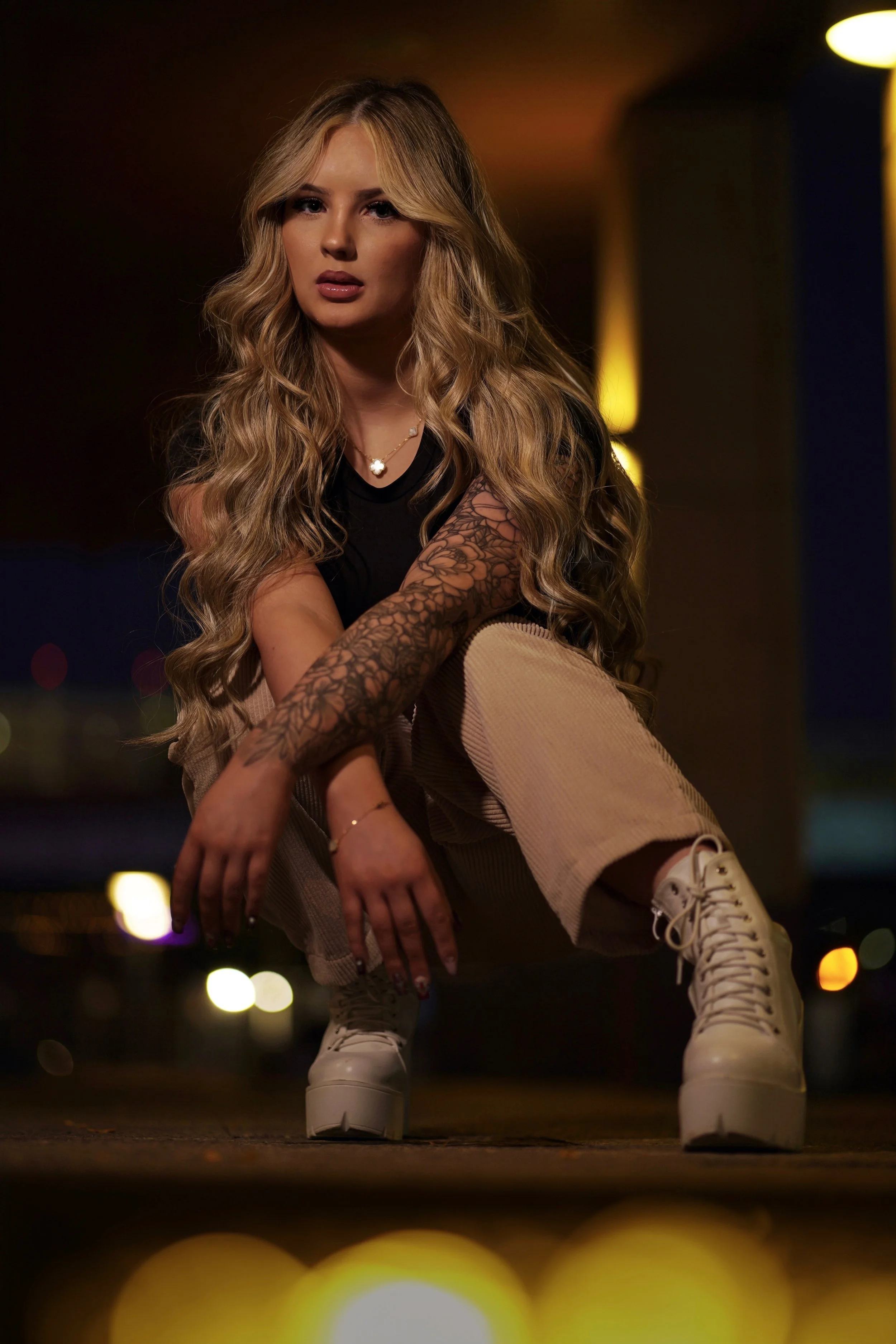 A woman with long, wavy blonde hair crouching in a dimly lit space at night, wearing a black t-shirt, beige pants, white platform boots, and displaying tattooed arms.