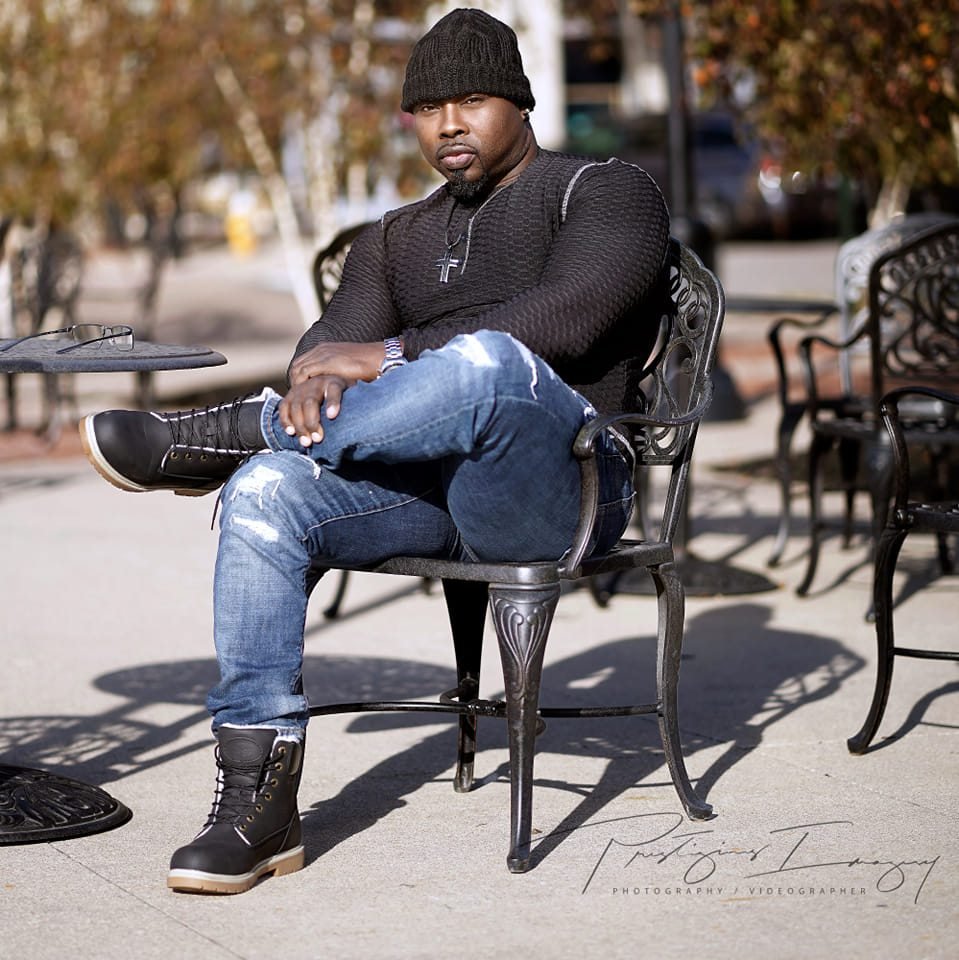 A man sitting on a black metal outdoor bench with his legs crossed, wearing a black knit hat, black sweater, ripped jeans, and black high-top boots, with a cross necklace, on a sidewalk in a park or plaza with trees and other chairs in the background.