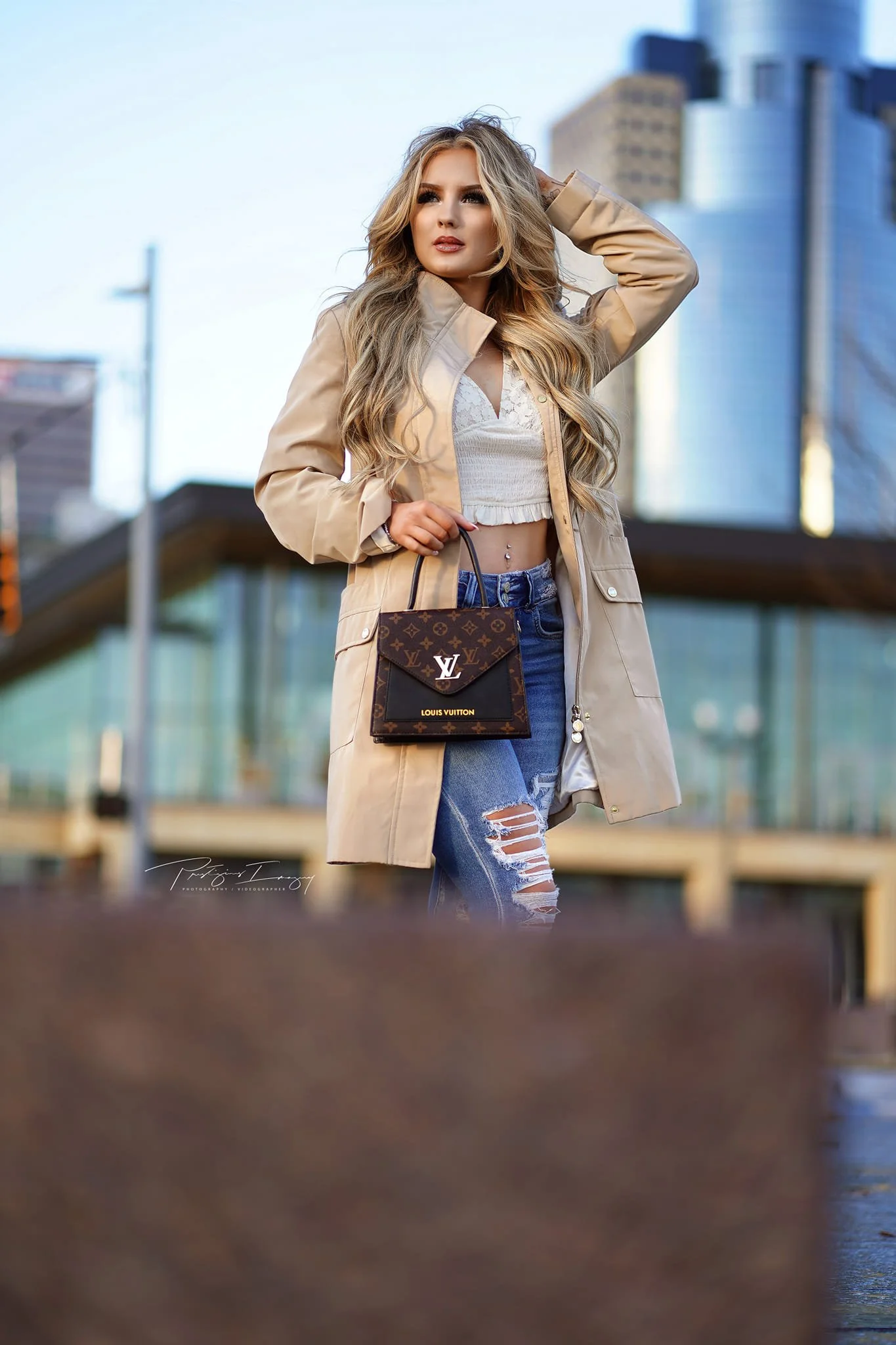 Fashionably dressed woman with long blonde hair, wearing a beige trench coat, white crop top, and ripped jeans, holding a Louis Vuitton handbag, standing outdoors in an urban setting.