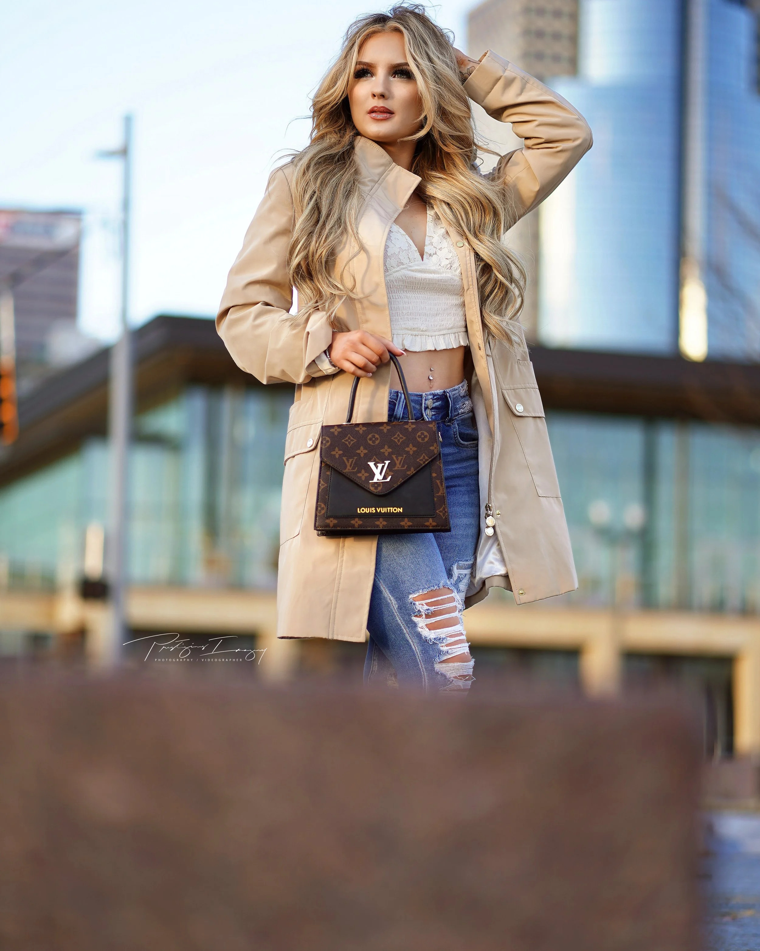 A woman in a tan coat, white crop top, and ripped jeans holding a Louis Vuitton handbag outdoors in an urban setting.