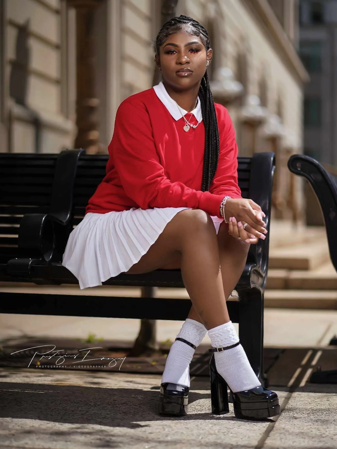 A young woman sitting on a black park bench outdoors, wearing a red sweater, white pleated skirt, and black platform shoes with white socks, with braided hair and jewelry, in an urban setting.