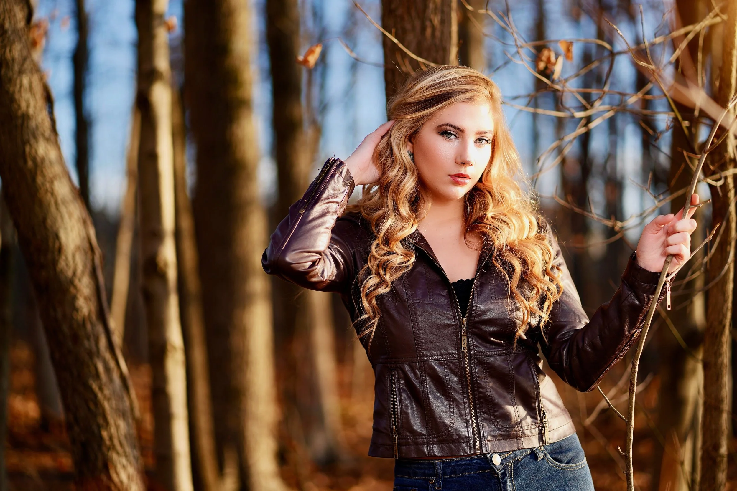 A young woman with long, curly blonde hair wearing a brown leather jacket and blue jeans, standing in a wooded area during fall, holding a thin tree branch.