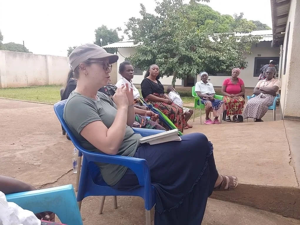 A group of women sitting outdoors on plastic chairs in a semi-circle, engaged in a discussion or meeting.