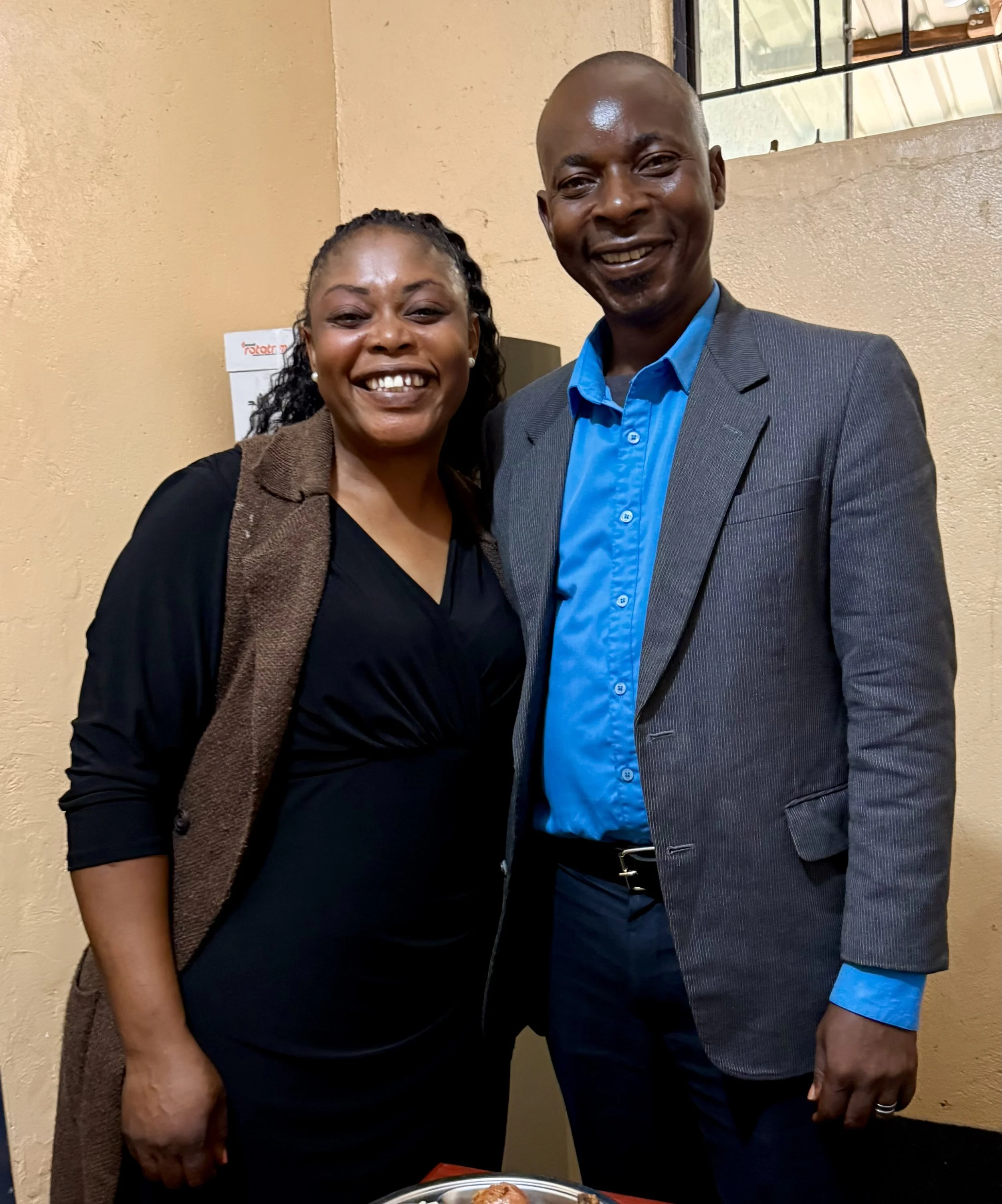 A smiling woman with curly hair and a brown scarf standing next to a man in a blue shirt and gray blazer, both smiling and posing for a photo indoors.