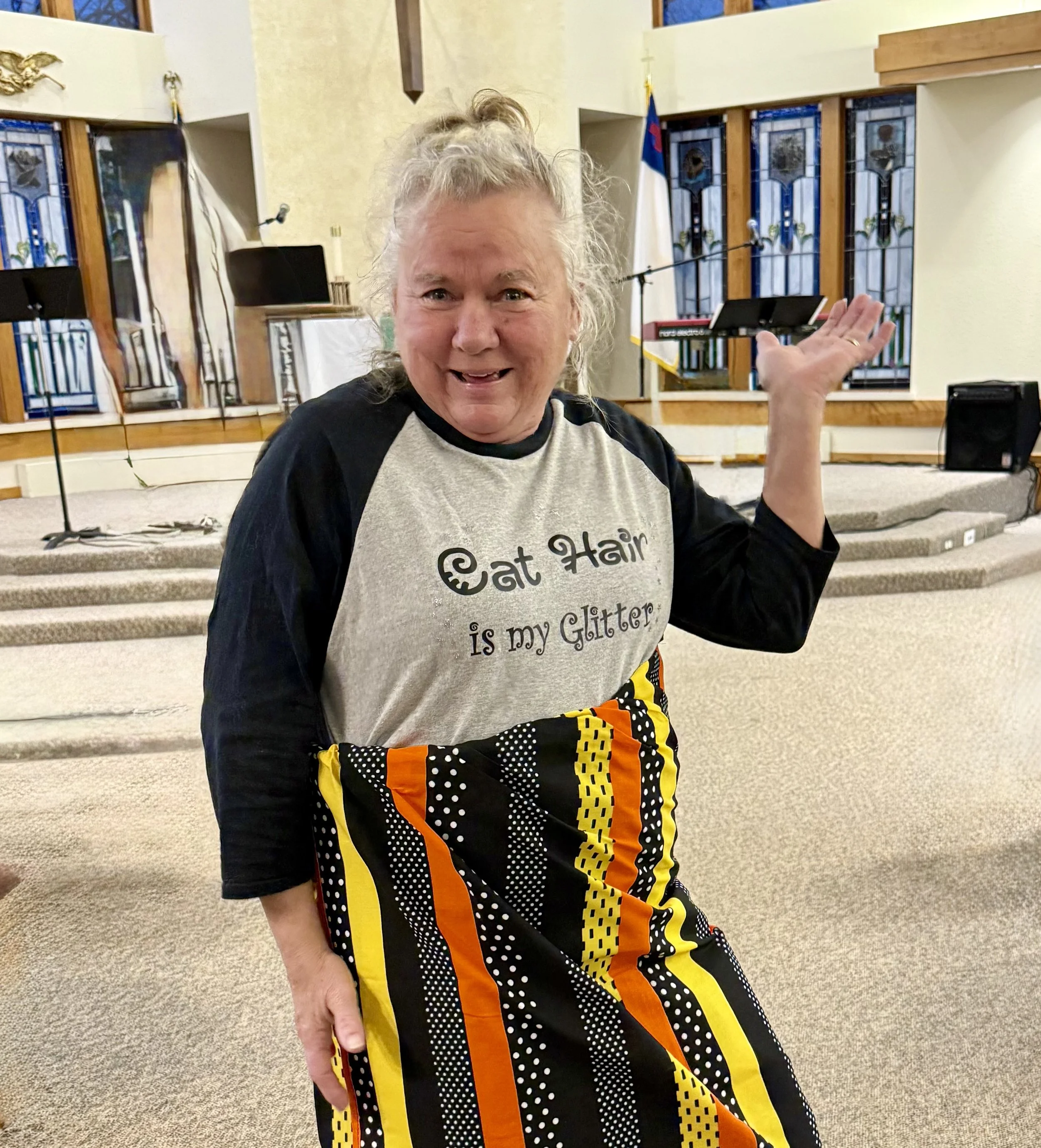 A smiling woman with gray hair styled in a messy bun, wearing a beige and black T-shirt that says 'Eat Hair is my Glitter' and colorful, patterned pants, standing inside a church with stained glass windows and a flag in the background.