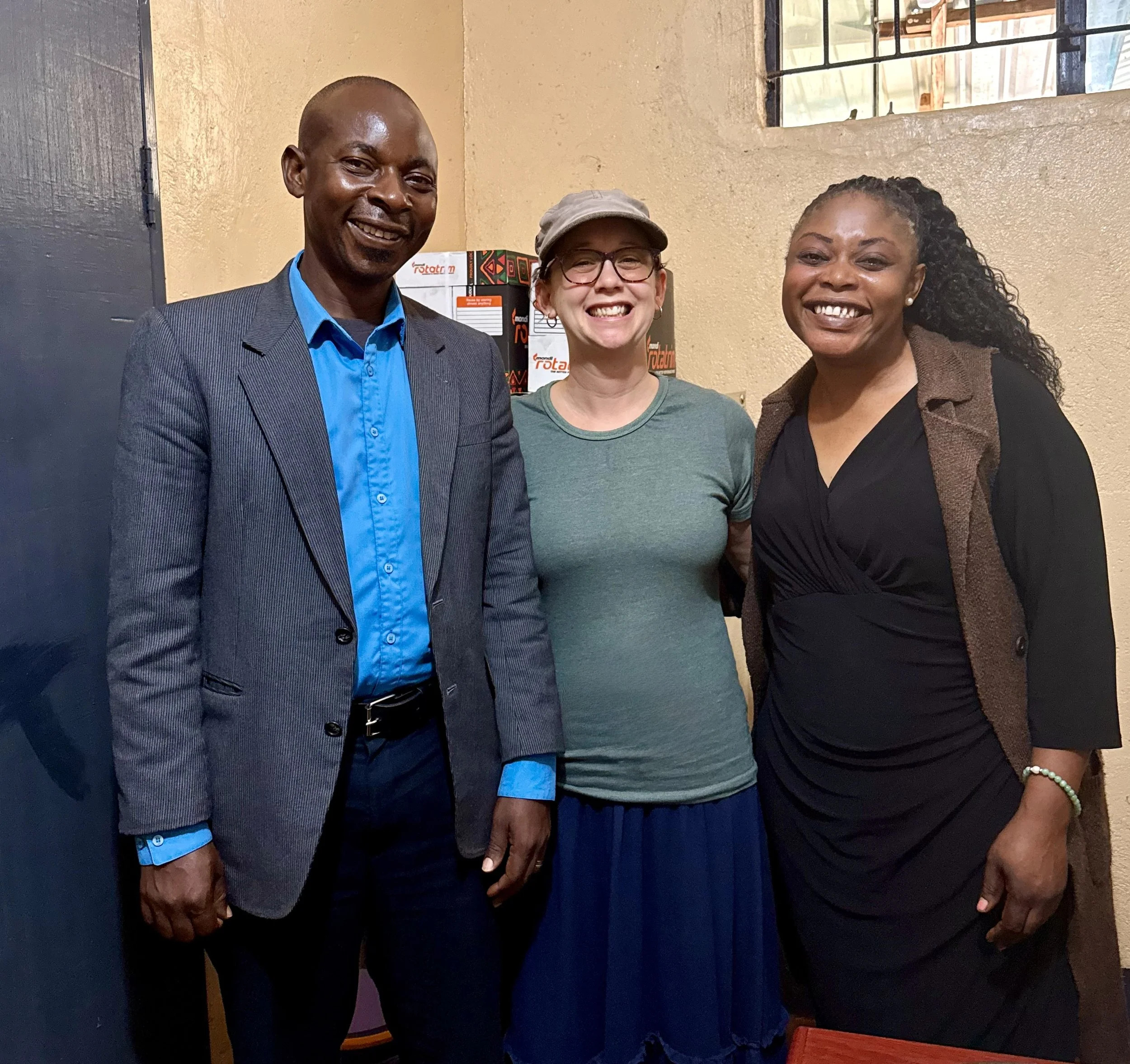 Three smiling women standing indoors near a beige wall and a barred window, dressed in casual and business attire.