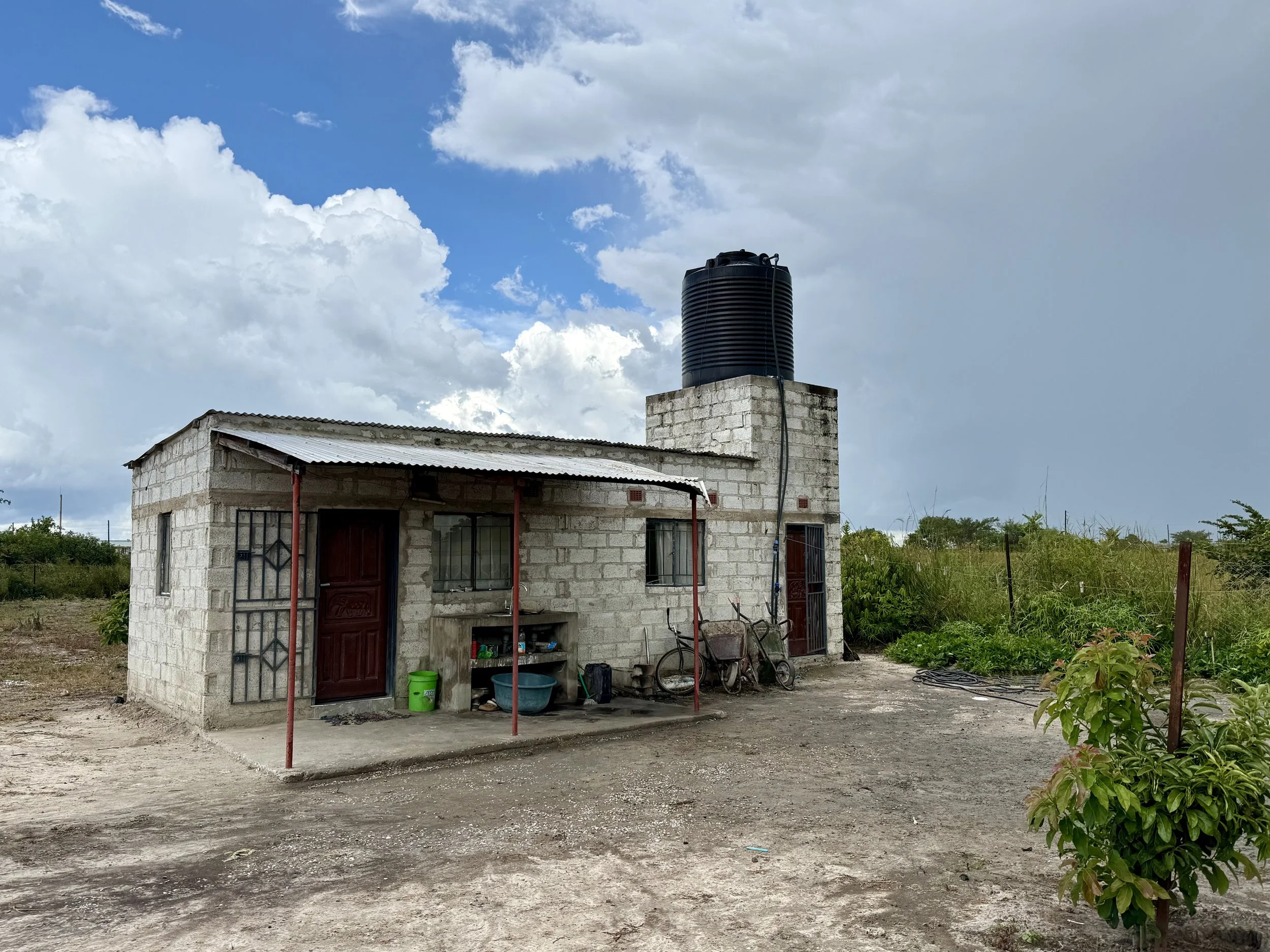 A small, unfinished house with a metal roof, two front doors, and a chimney on a concrete block. The house has various tools and buckets outside. A black water tank is on top of the chimney. The surroundings include dirt yard and green plants, with a cloudy sky overhead.