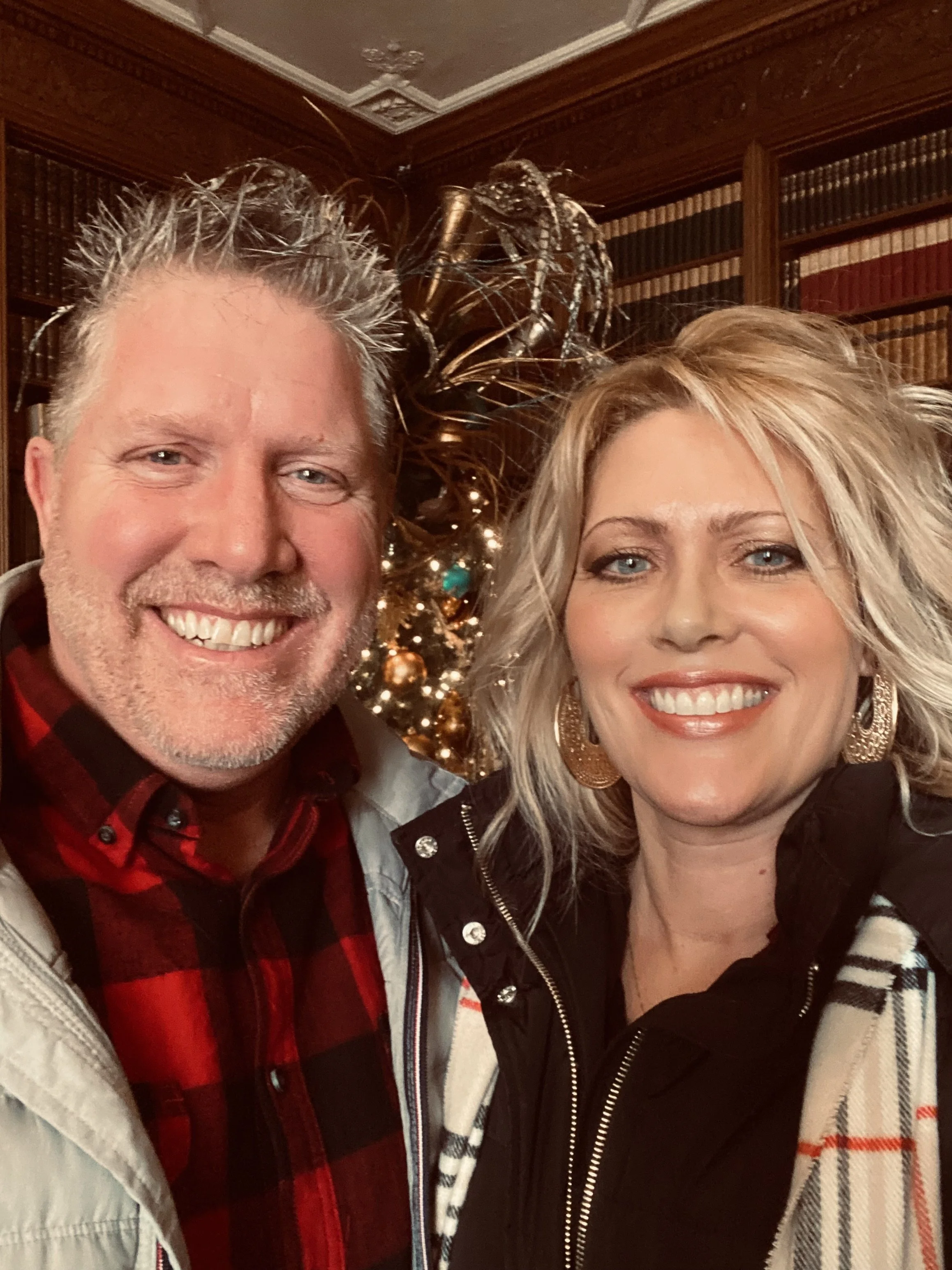 A man and woman smiling for a selfie indoors, with a decorated Christmas tree and a ornate wood bookshelf in the background.