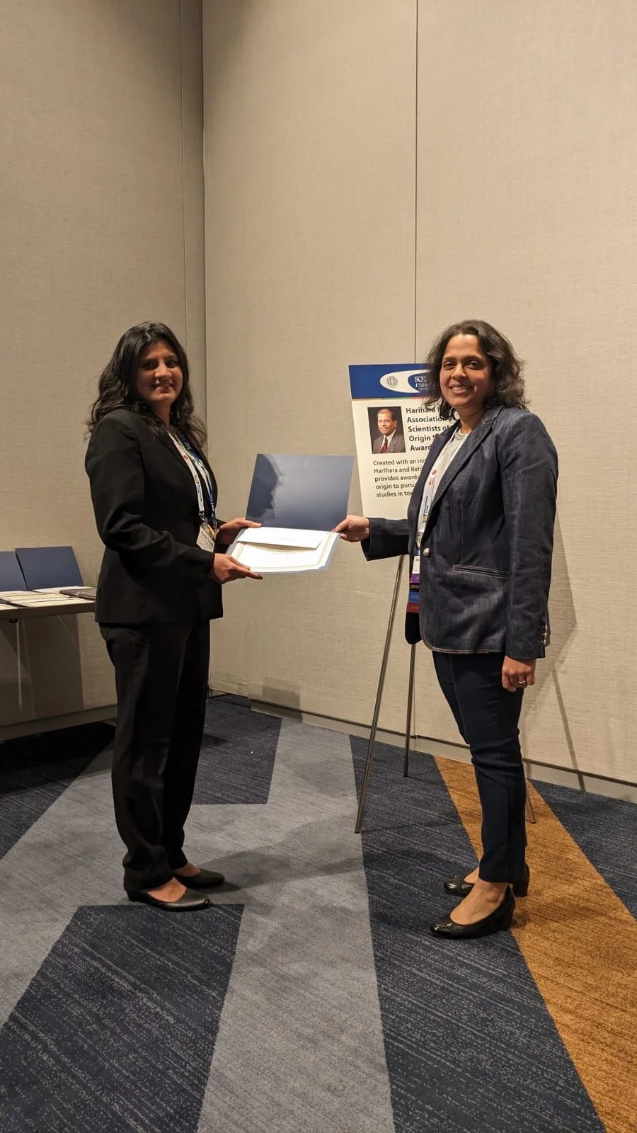 Two women at an award ceremony; one woman is presenting an award or certificate to the other, standing next to a poster board with a photo and description.
