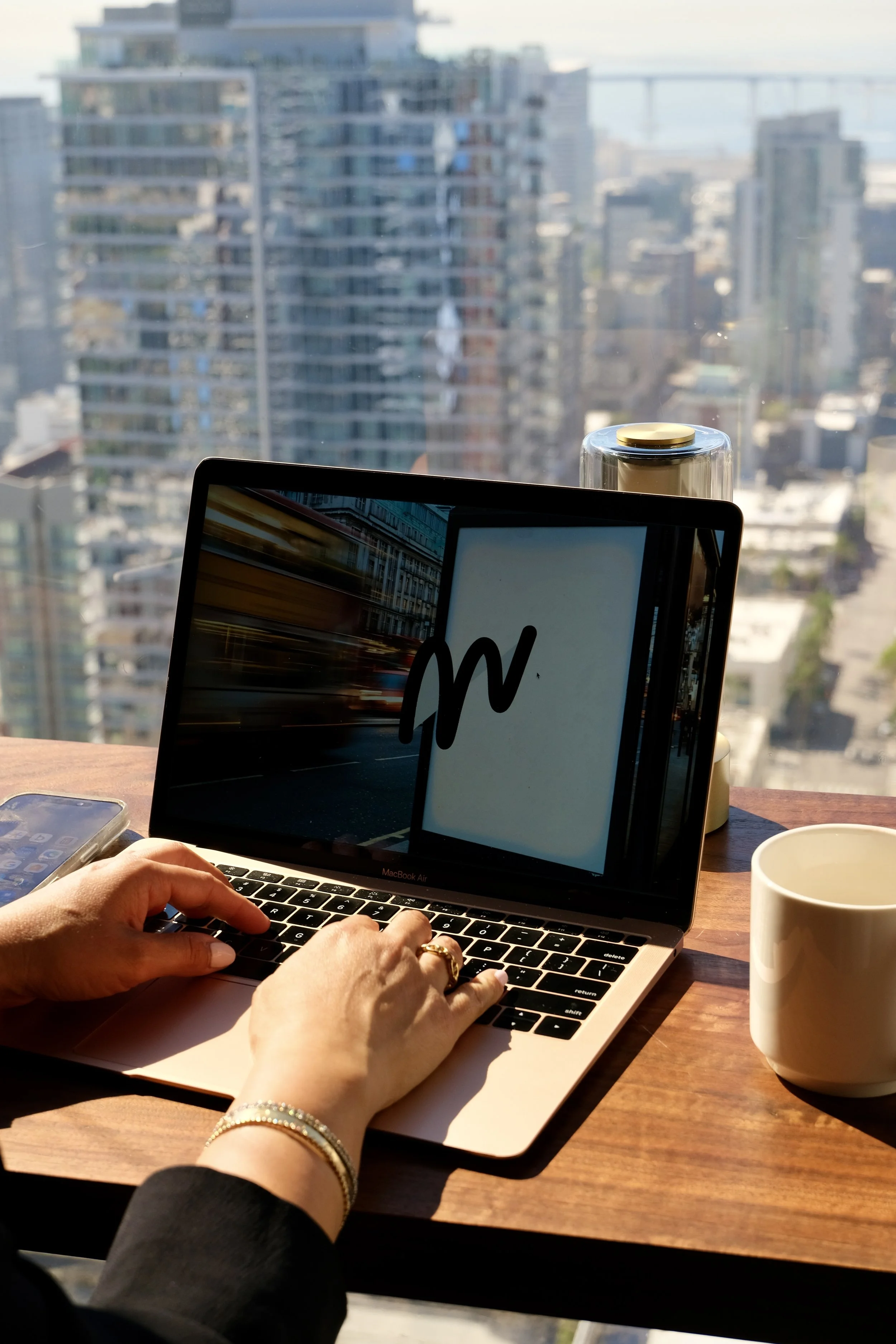 A person types on a MacBook Air laptop at a wooden desk with a city skyline view in the background. A cup and a glass container are also on the desk.