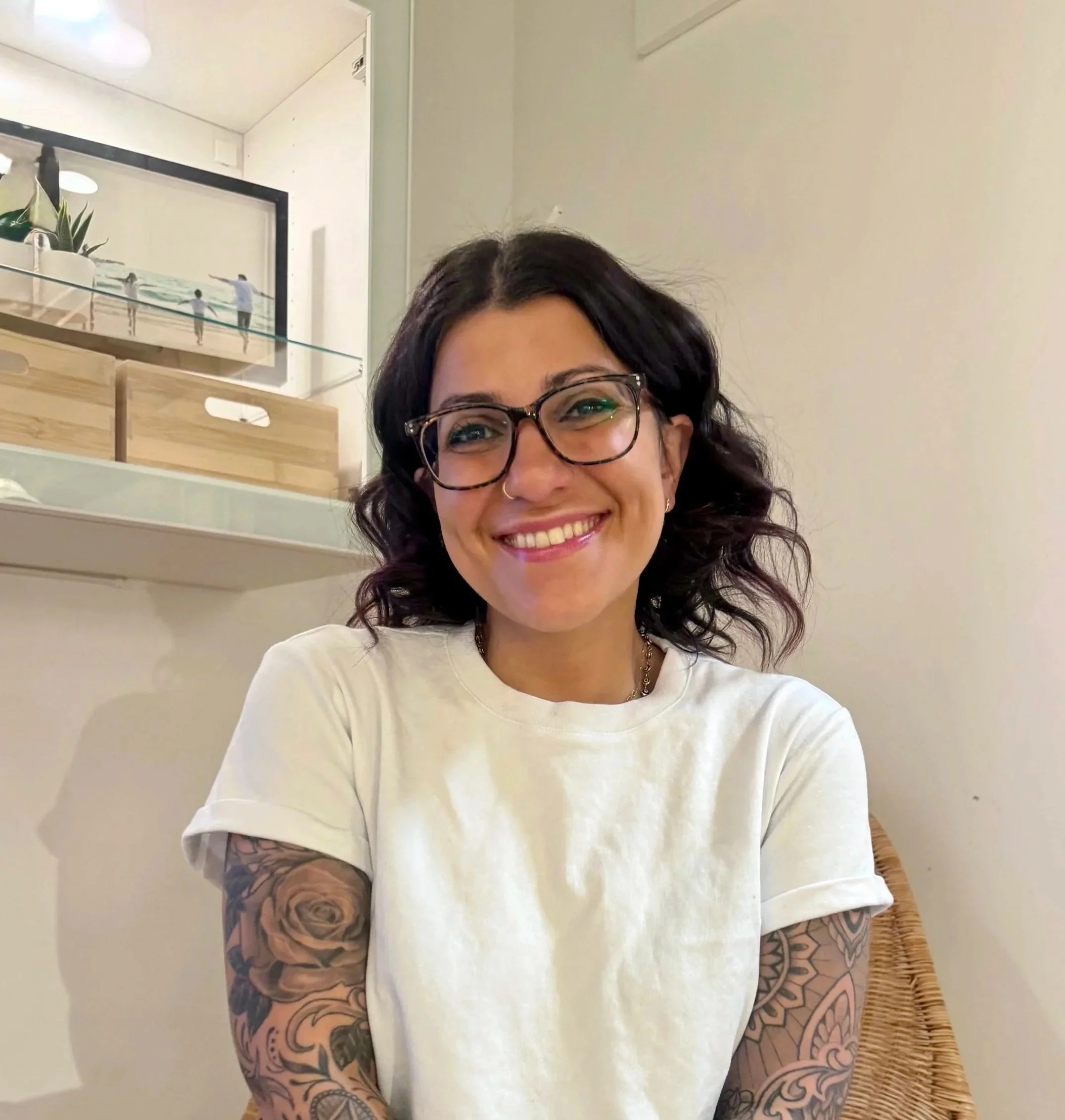Smiling woman with glasses and dark wavy hair, wearing a white t-shirt, tattooed arms, in a room with decorated walls and a wicker chair.