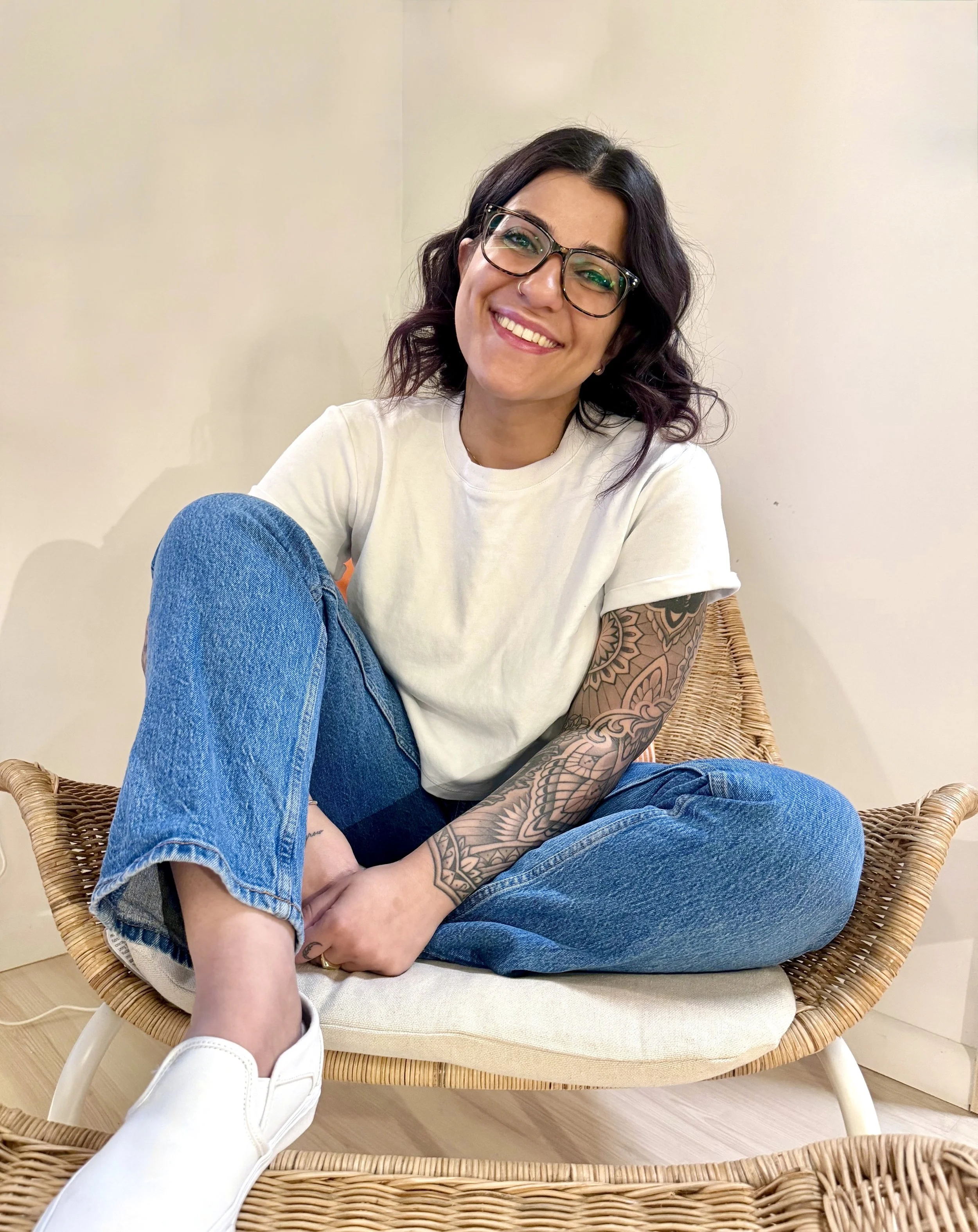 A young woman with short black hair, glasses, and a nose ring smiling while sitting cross-legged in a wicker chair, wearing a white T-shirt and blue jeans, with an arm tattoo visible, in a room with light-colored walls.