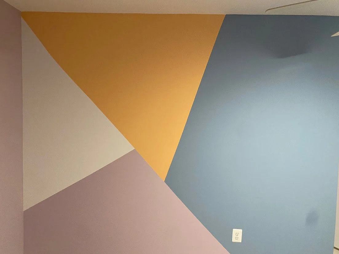 Modern three-tone geometric mural in mustard, slate blue, and blush pink on a corner accent wall