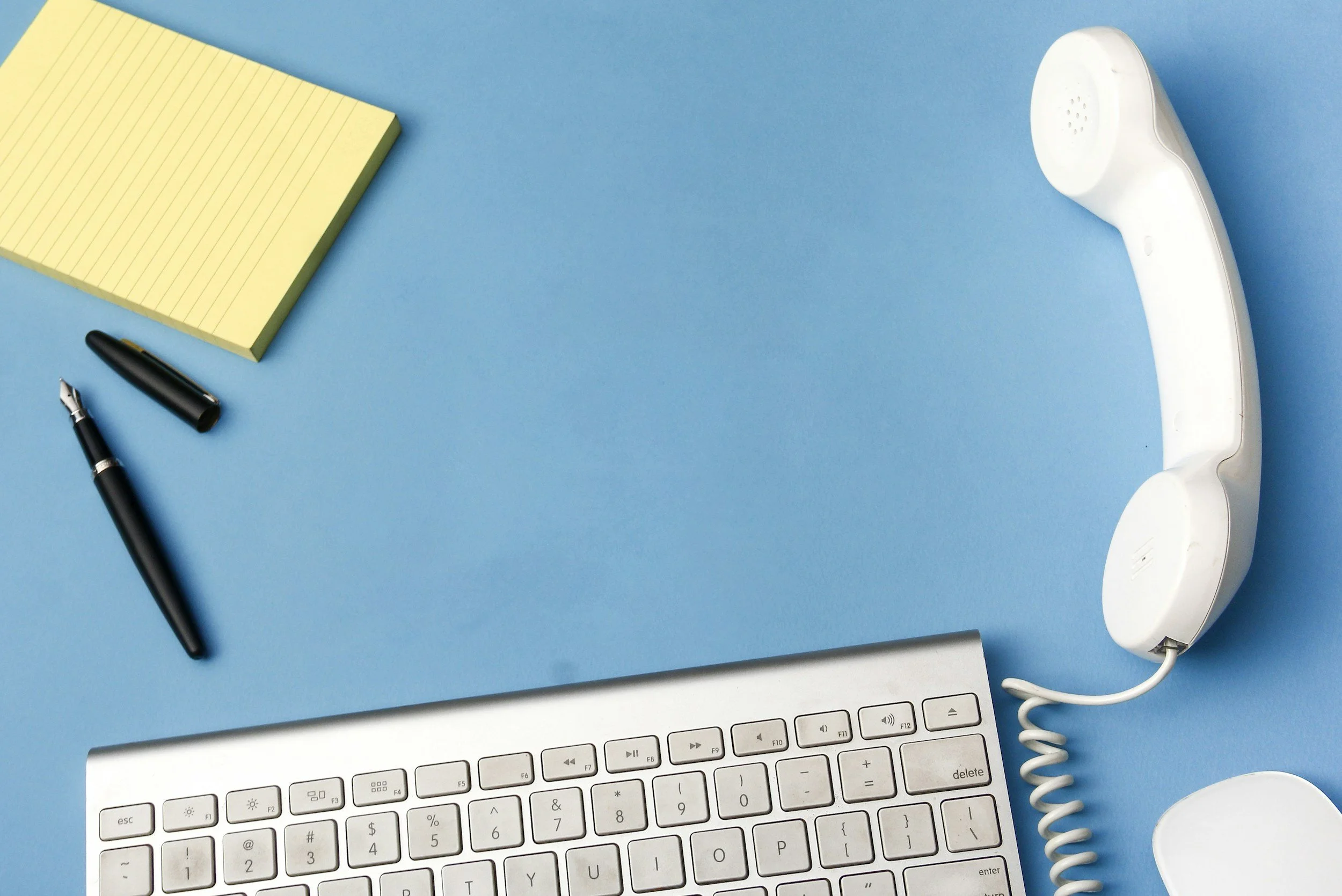 Office desk setup with a white telephone, a white keyboard, a black pen with the cap removed, a small yellow notepad, and a white circular object on a light blue background.