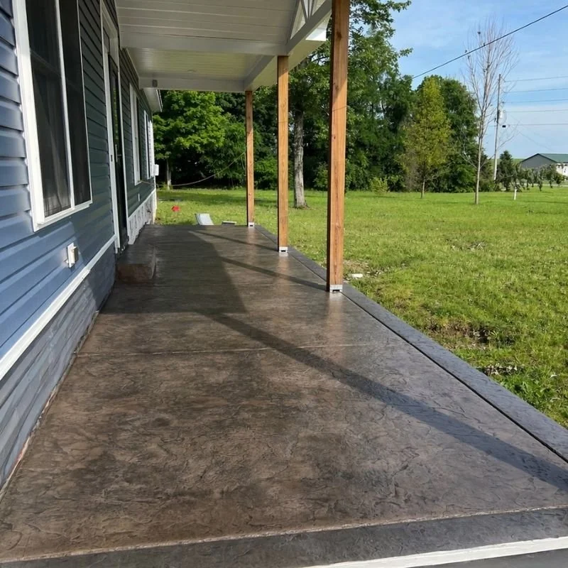Newly poured concrete patio with wooden porch posts on a house with blue siding, in a green grassy yard with trees and clear blue sky.