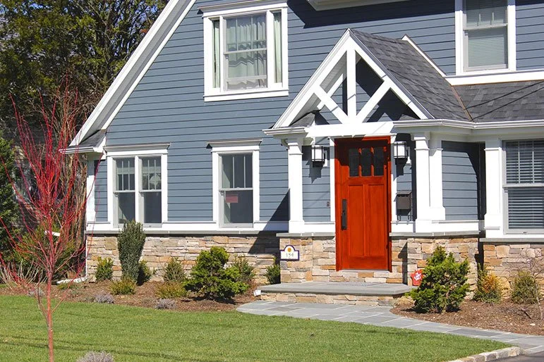 Front of a blue house with a red wooden door, white trim, and a stone foundation. There are neatly landscaped bushes and a small tree in the yard.