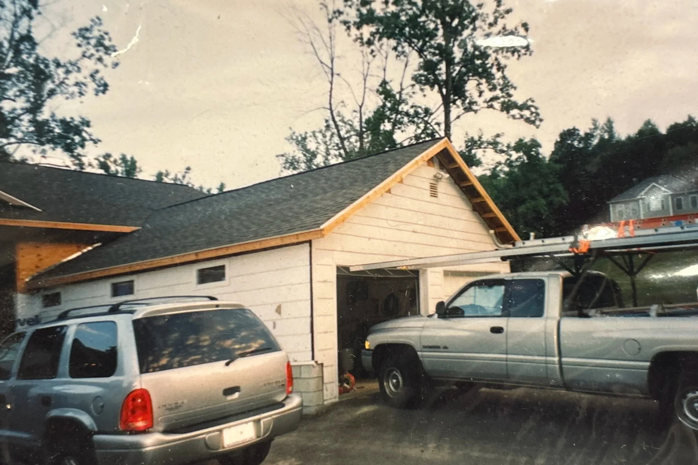 A house under construction with a partially finished roof and siding, two parked vehicles in front, and trees in the background.