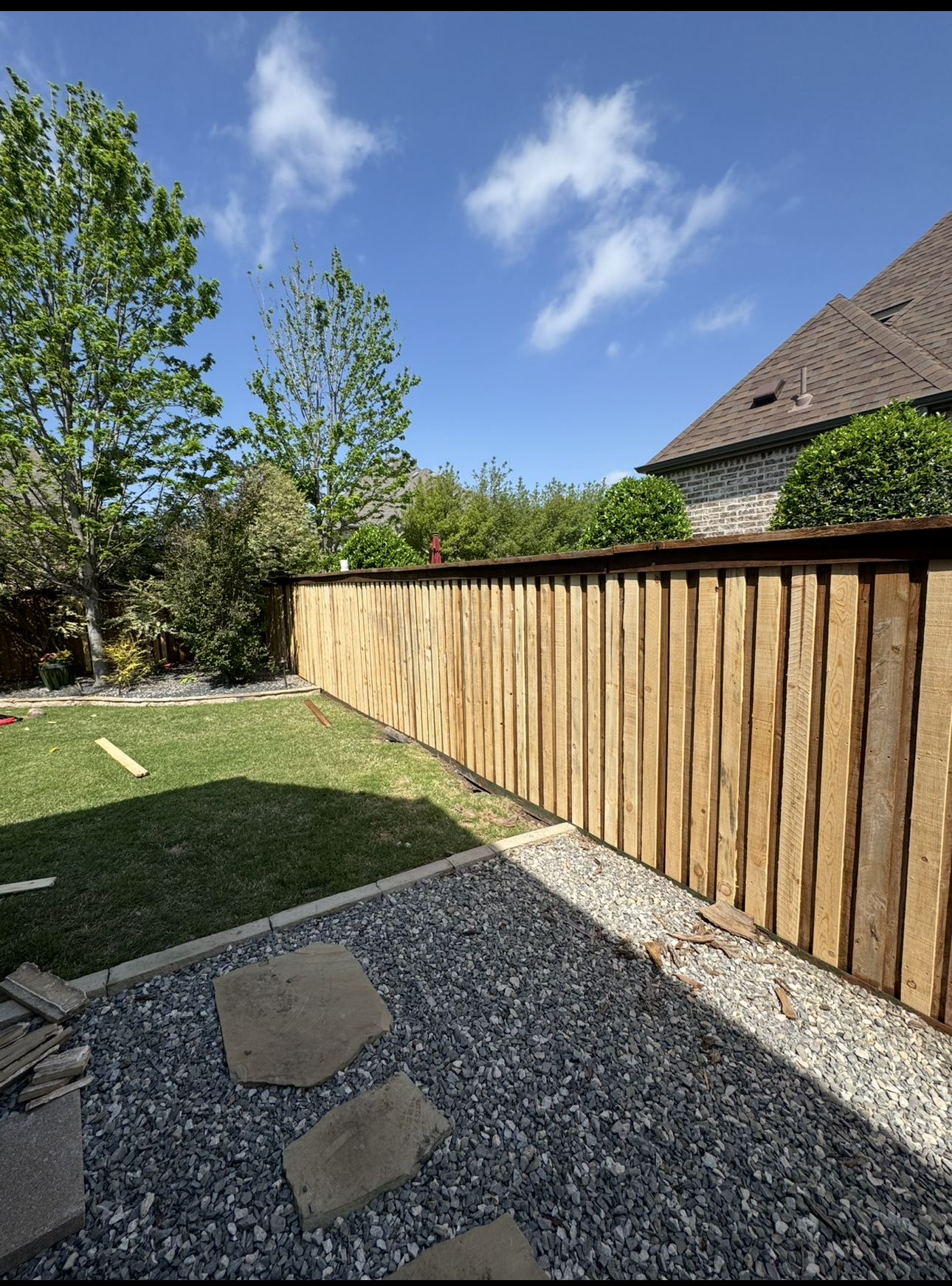 A backyard with a newly built wooden fence, green grass, trees, and a brick house with a shingled roof under a blue sky with scattered white clouds.