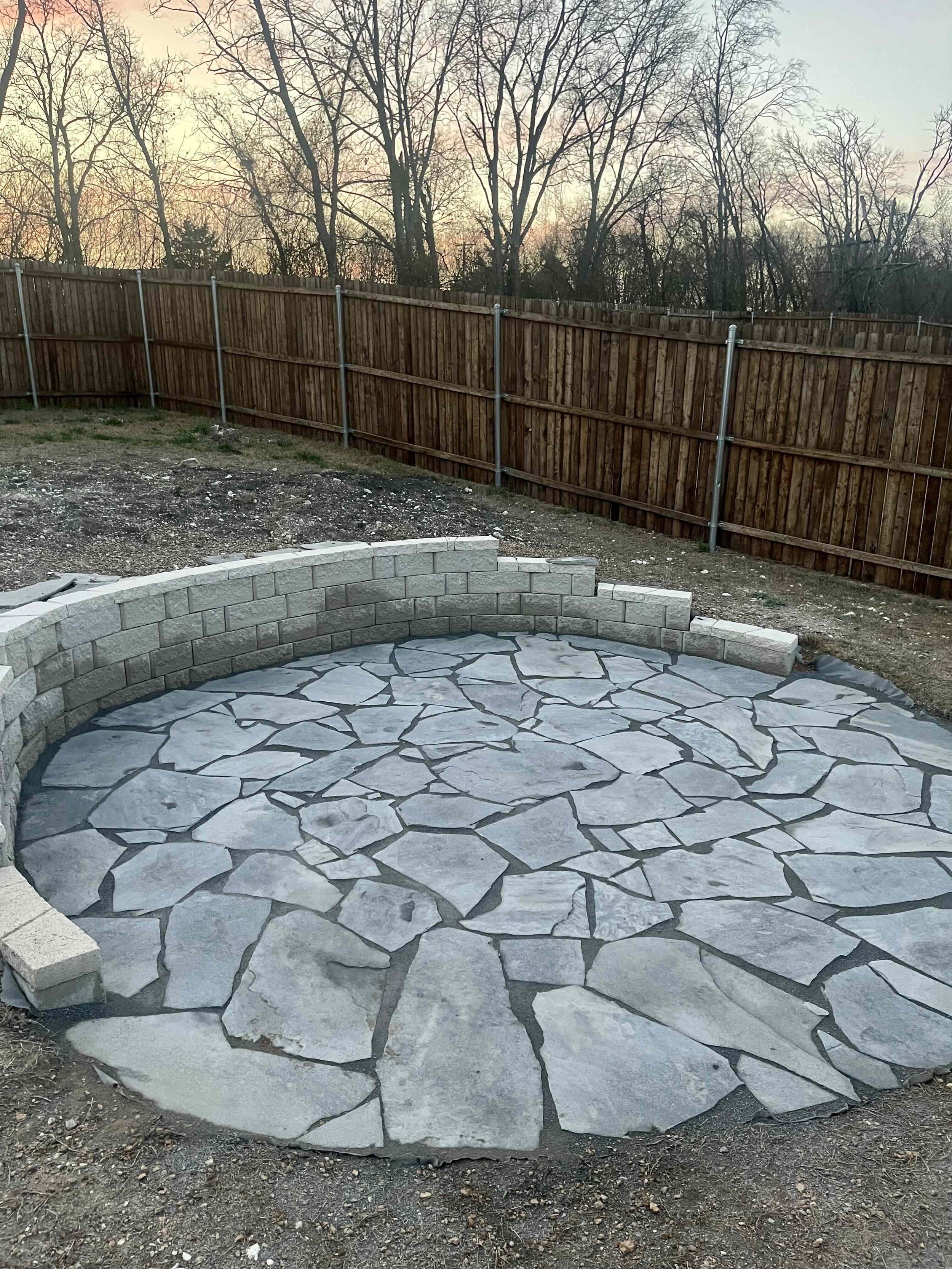A backyard patio with a circular stone surface, a semi-circular brick wall, a wooden fence, and leafless trees in the background during sunset.