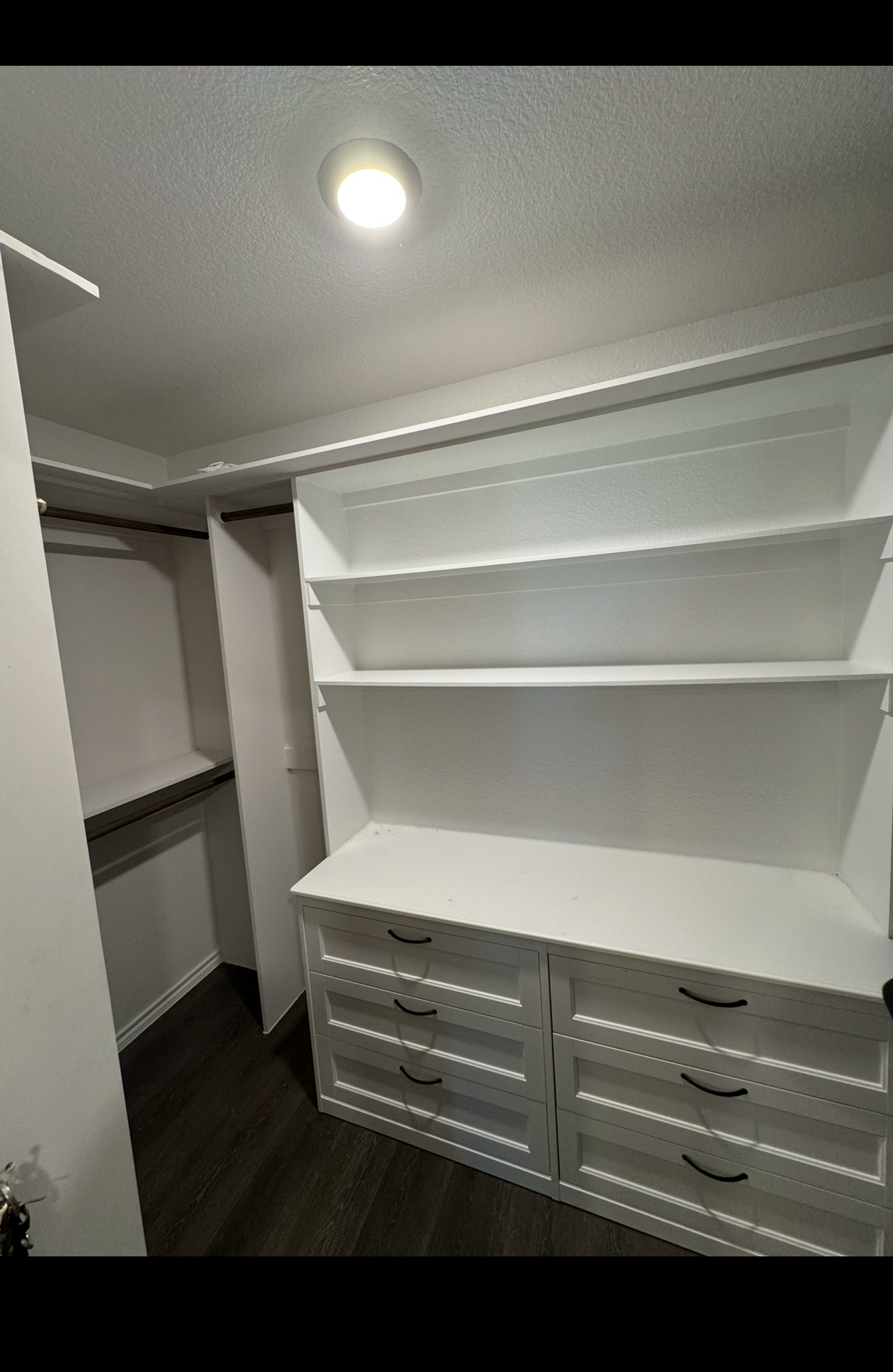 Empty walk-in closet with white built-in shelves and drawers, dark wood flooring, and a ceiling light.
