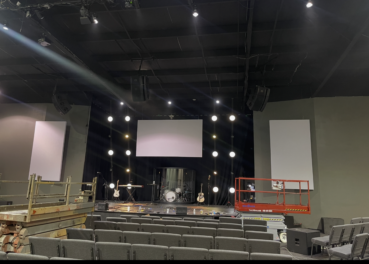 An empty auditorium or concert hall with a stage setup for a live performance, including musical instruments such as guitars, a drum set, and microphones, along with seating and construction equipment like a scissor lift.