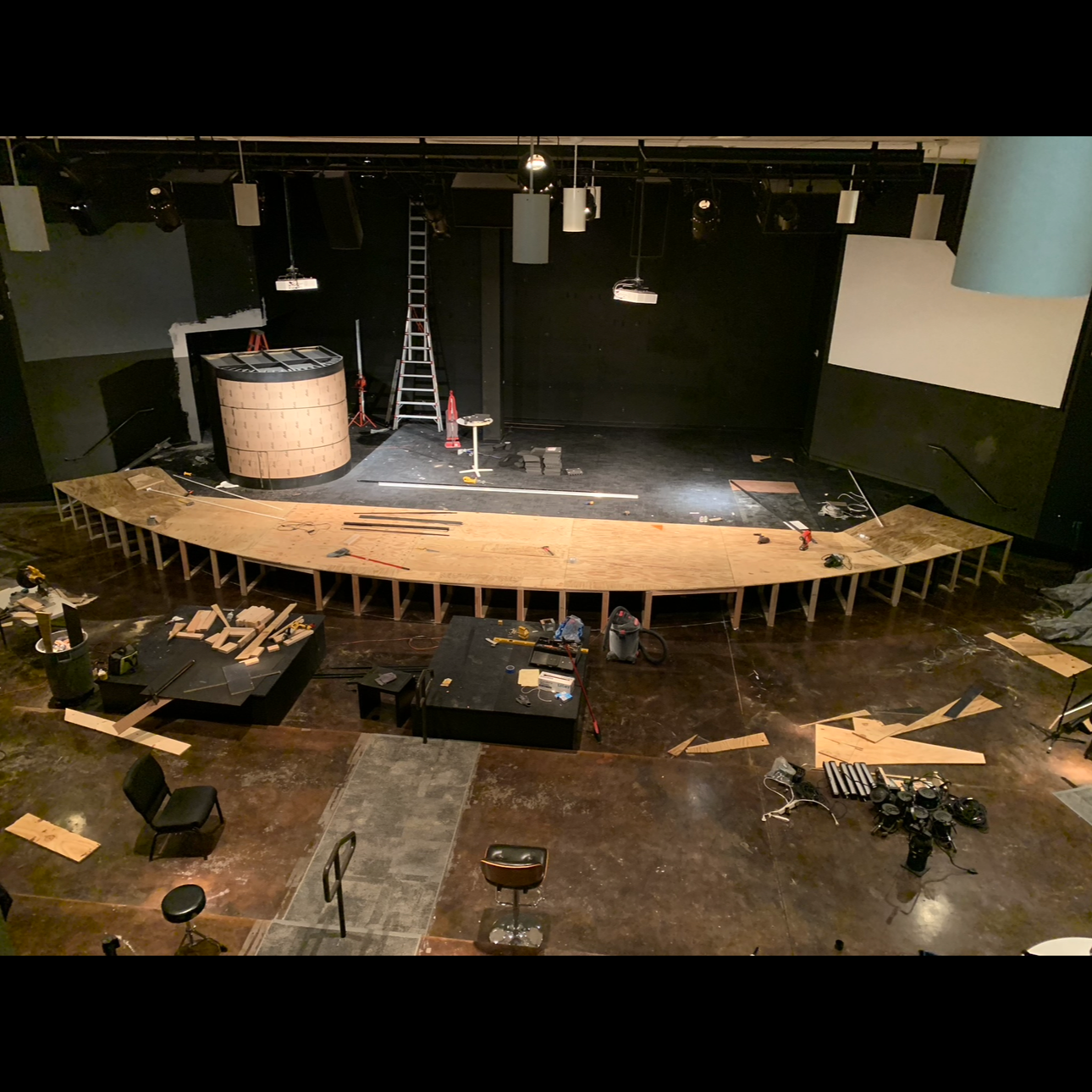 Stage under construction with wooden platform, ladder, tools, and chairs. Black walls, ceiling speakers, and lighting fixtures.