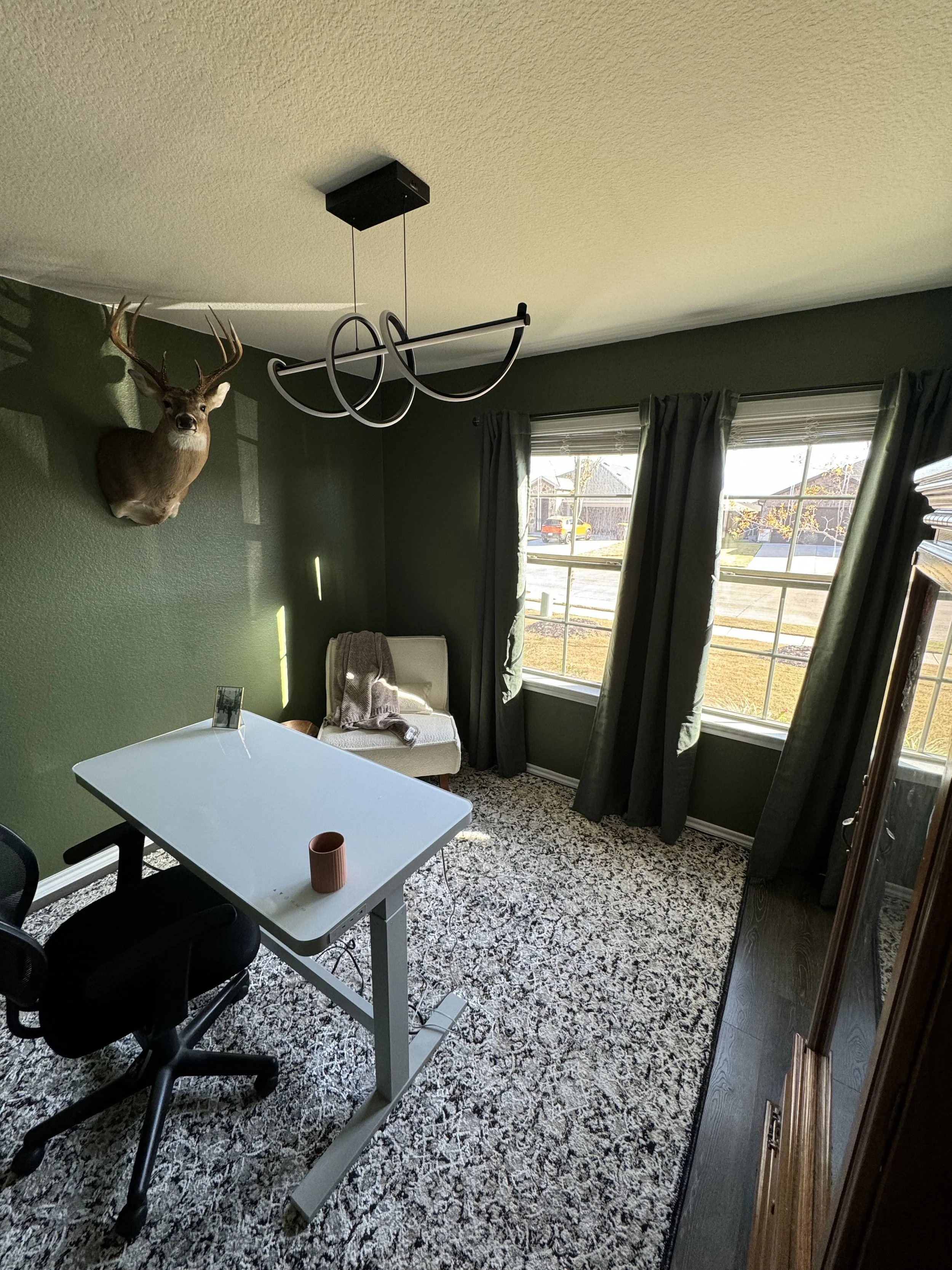 A room with dark green walls, a white desk, a black office chair, a beige armchair with a towel on it, and windows with dark curtains. There is a mounted deer head on the wall and a modern black chandelier hanging from the ceiling.