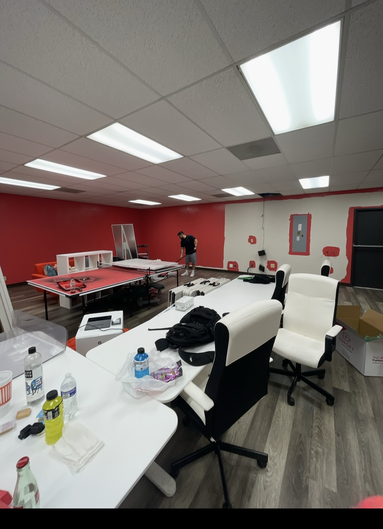 Office or conference room under renovation with a worker painting a wall red, several tables with items and chairs, and a large ceiling with fluorescent lights.