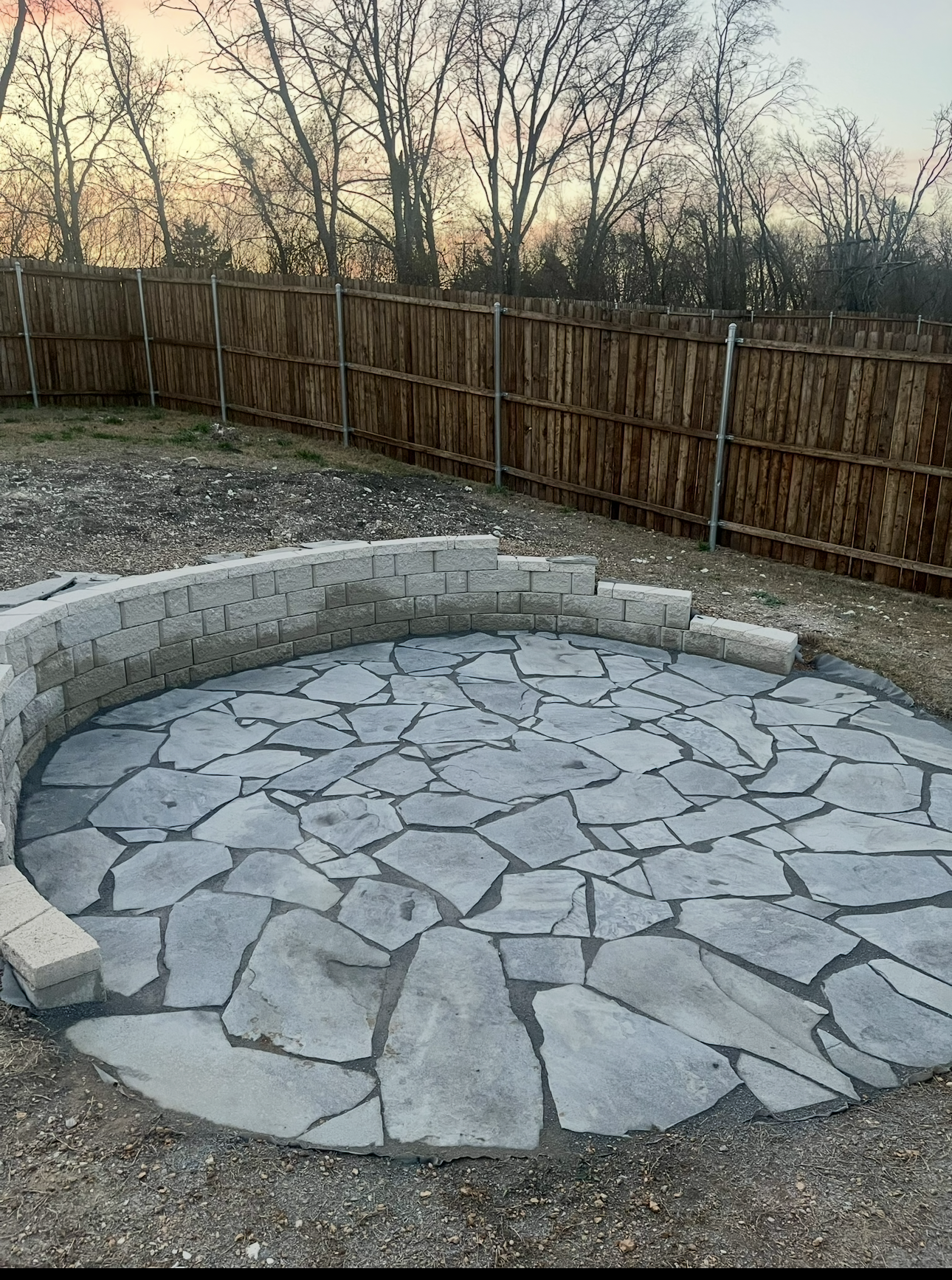 A backyard with a newly installed stone patio and a curved wooden fence. The patio features large, irregularly shaped gray stones set in mortar. In the background, there are leafless trees and a sunset sky.