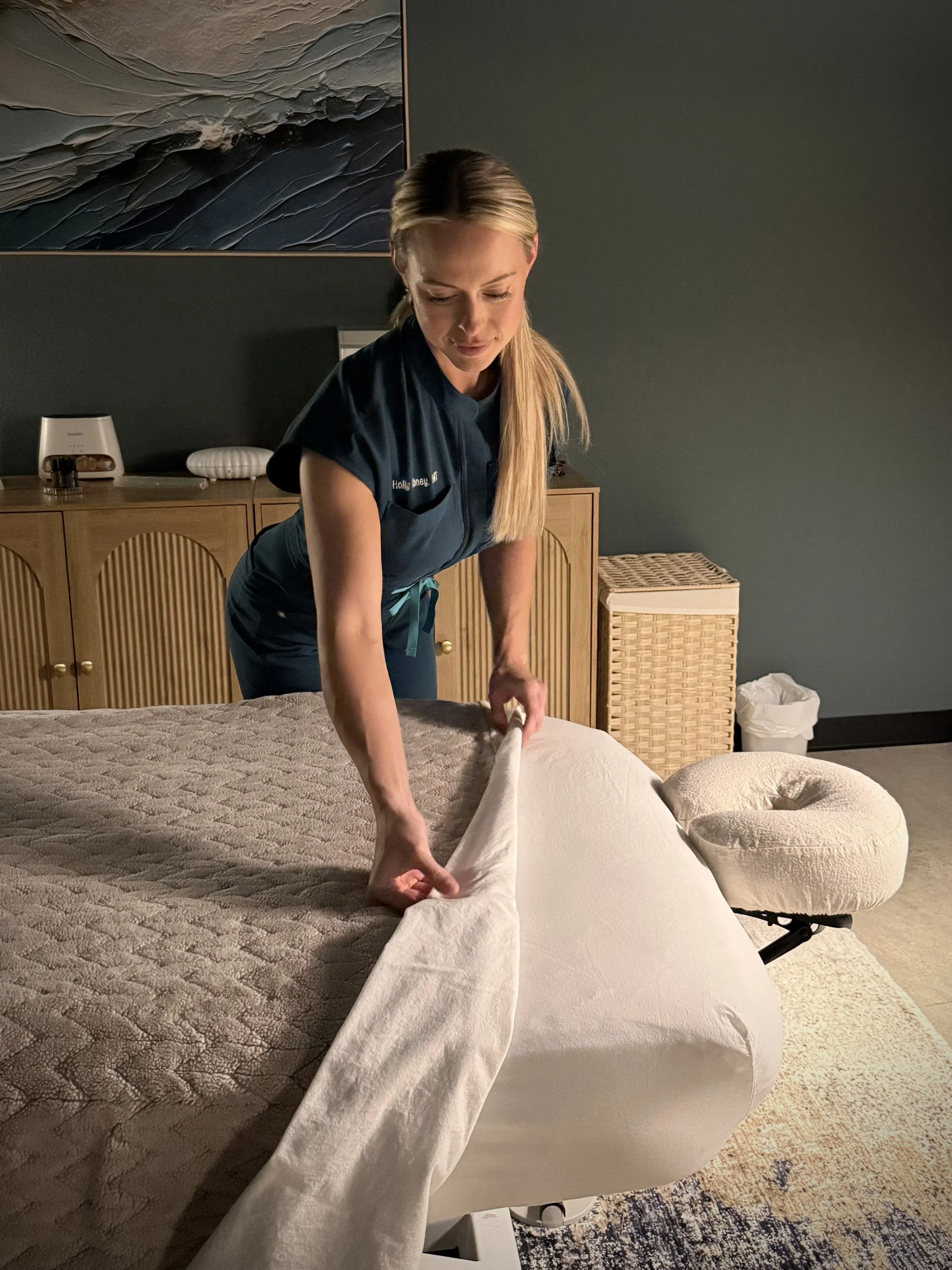 A woman making the bed in a bedroom, arranging a white duvet cover on a bed with a brown quilted bedspread. She has long blonde hair and is wearing a dark blue shirt.