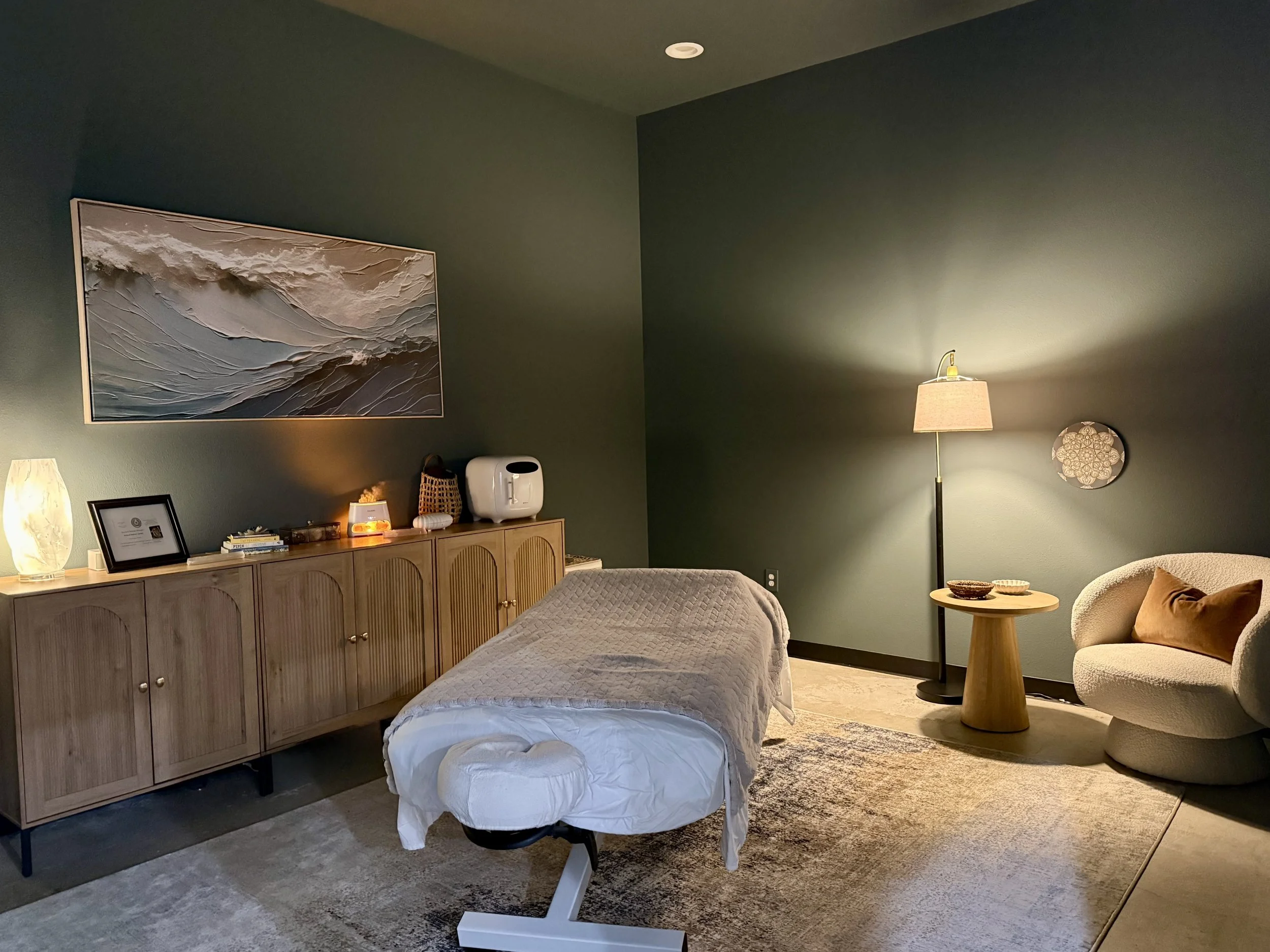 A cozy bedroom with a green wall, a large ocean-themed wall art, a wooden cabinet with small decorative items, a white bed, a beige armchair with a brown cushion, a wooden side table with bowls, a floor lamp with a beige shade, and a small white humidifier.