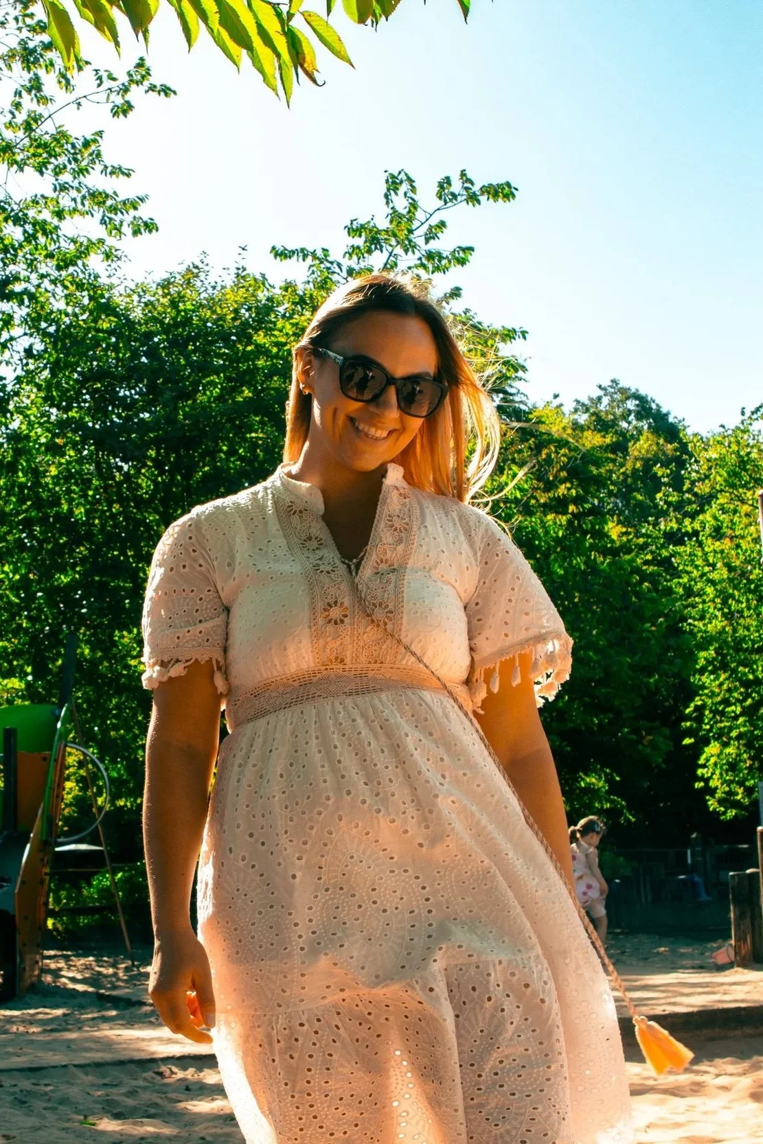 Smiling woman wearing sunglasses and a white Eyelet dress standing outdoors in a park with trees and playground in the background