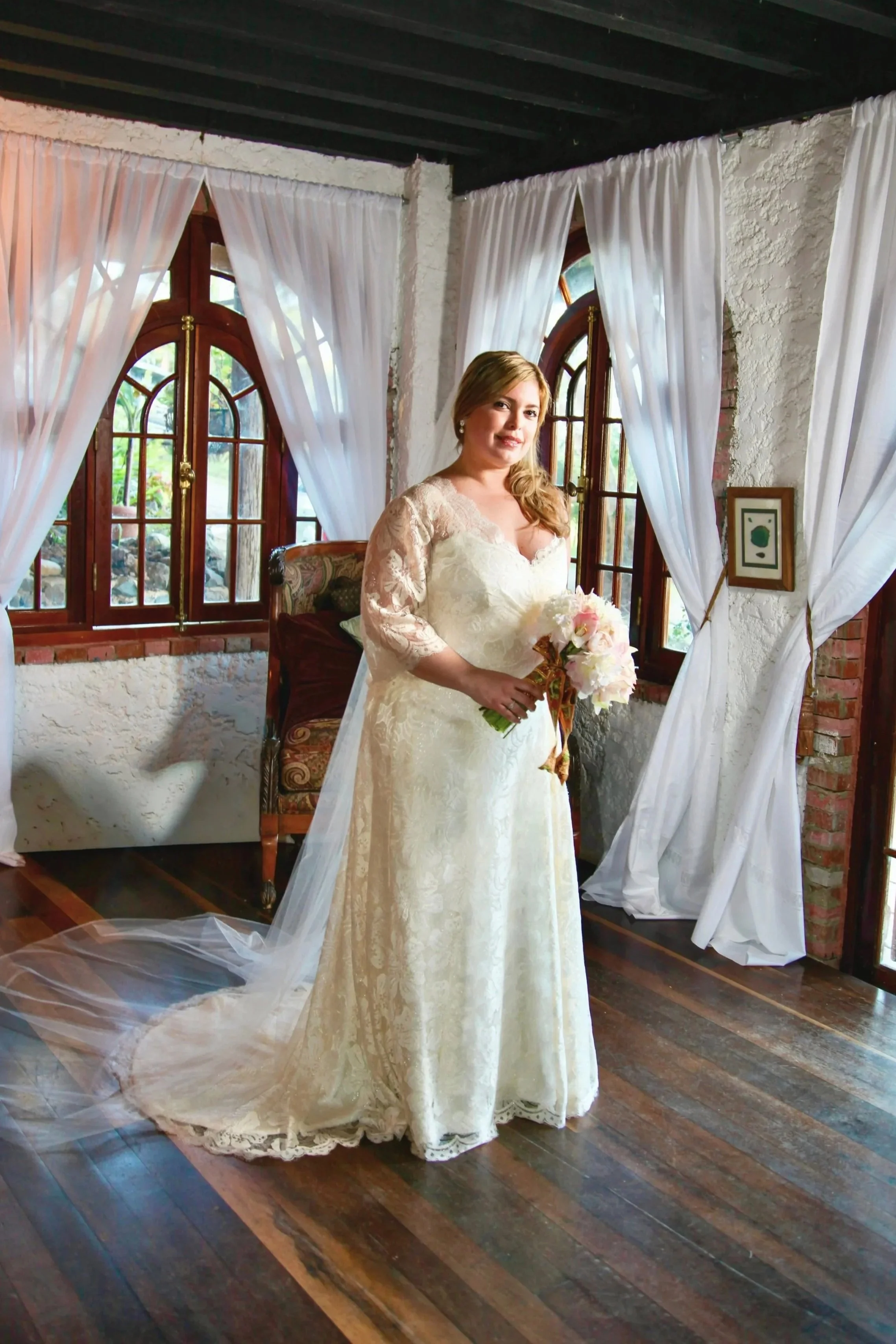 A plus size bride, a woman, Mariela Resta, in a white wedding dress standing indoors with wooden floors, white brick walls, large arched windows with curtains, holding a bouquet of pink and white flowers.