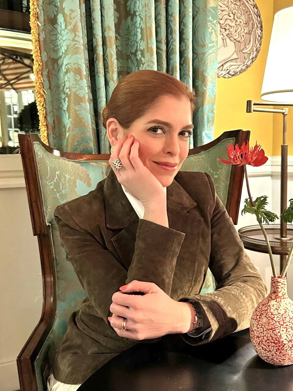 A woman, Mariela Resta, with auburn hair styled back, wearing a brown suede jacket and multiple rings, sitting at a table with a red and white patterned vase holding a red flower, in a warmly lit room with colorful curtains and a lamp.
