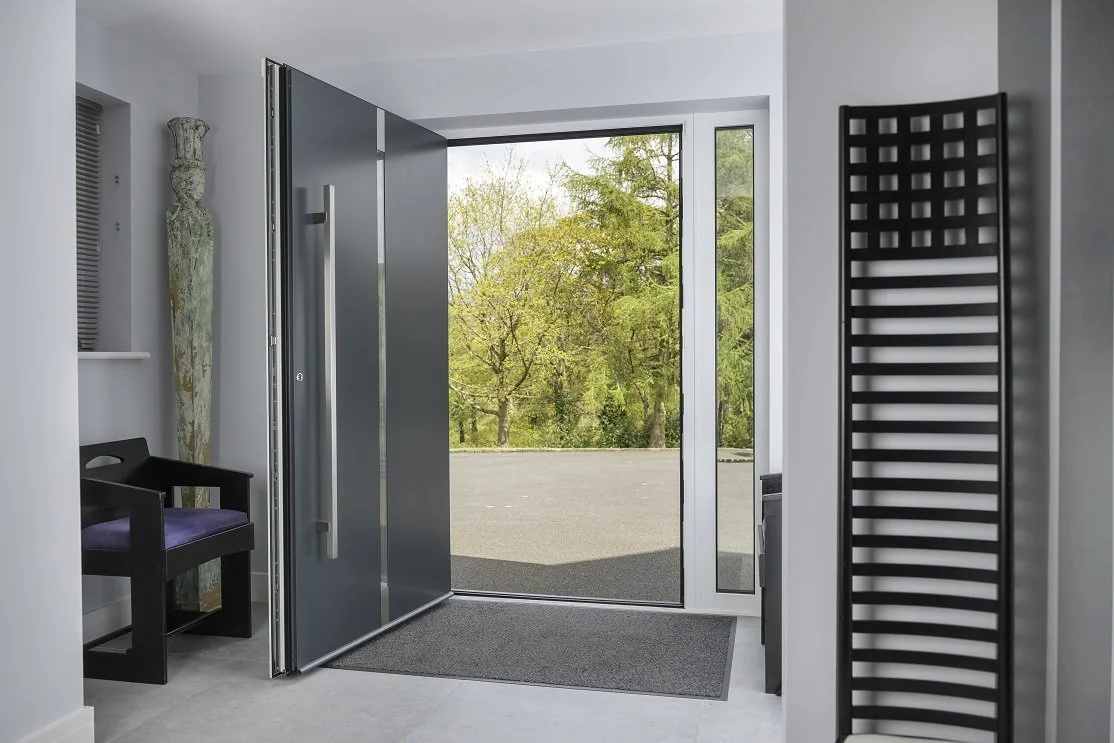 Open front door leading outdoors with trees and green foliage visible through glass, interior includes black chair with purple cushion, black heated towel rack, and an art piece on the wall.